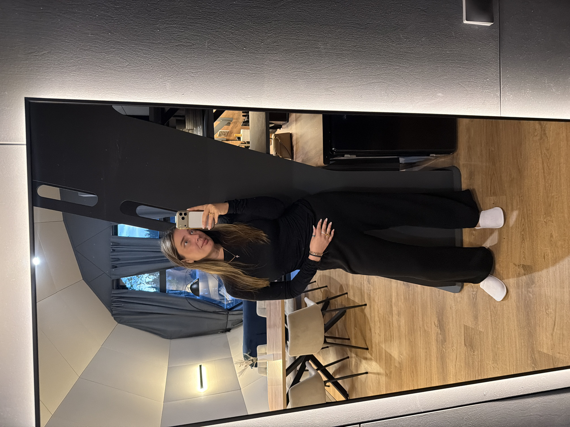 A woman is taking a mirror selfie in a modern apartment. She is wearing a black long-sleeved top and black wide-leg pants, paired with white slippers. The apartment features a wooden floor, a large mirror with integrated lighting, and a unique architectural ceiling. In the background, visible in the mirror's reflection, is a dining area with a wooden table and chairs, as well as a glimpse of a sofa and curtains. The overall aesthetic suggests a contemporary and minimalist living space. The city of Solov'evo, Russia is not depicted or indicated in the image.