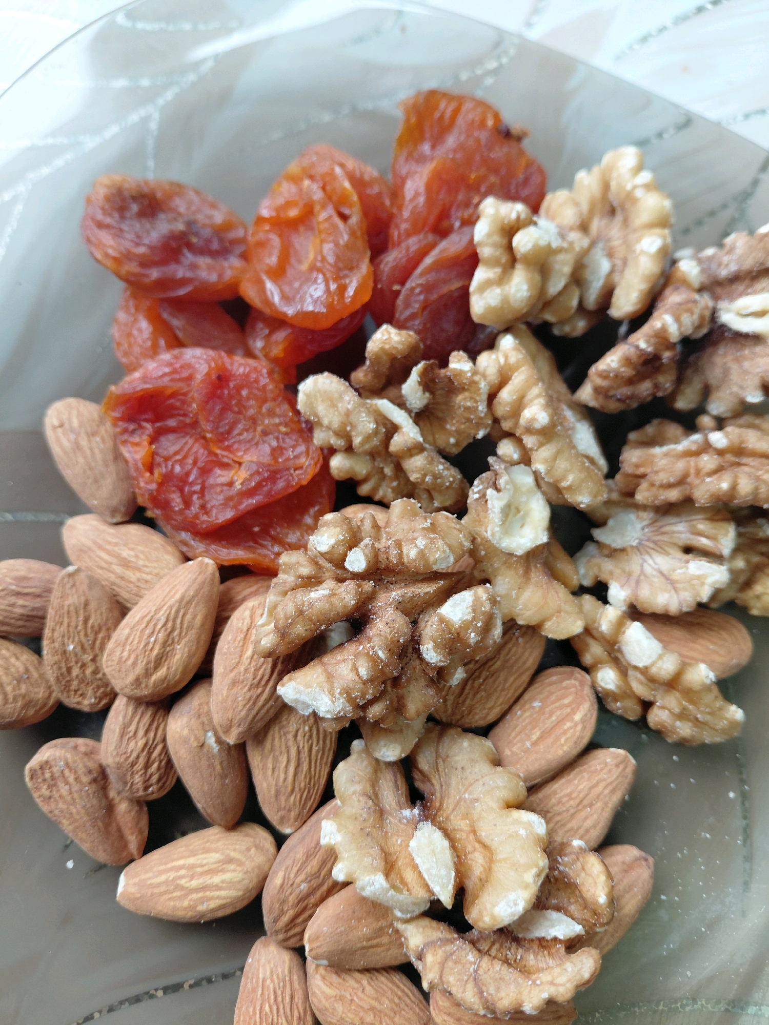 The image shows a close-up, overhead view of a mixture of dried apricots, walnuts, and almonds in a clear container or bowl. The dried apricots are a vibrant orange-red color and appear wrinkled and leathery. The walnuts are a light brown color and have their characteristic irregular, brain-like shape, with some showing remnants of white powdery residue. The almonds are oval-shaped with a light brown skin, and their surface has a slightly textured appearance. The items are piled together, with the almonds forming a base layer in the lower part of the image, the dried apricots clustered in the upper left, and the walnuts interspersed throughout. The background is out of focus, showing a light-colored, possibly patterned surface. There is no indication of the city of Malinovka, Russia, or any context related to it within the image itself.