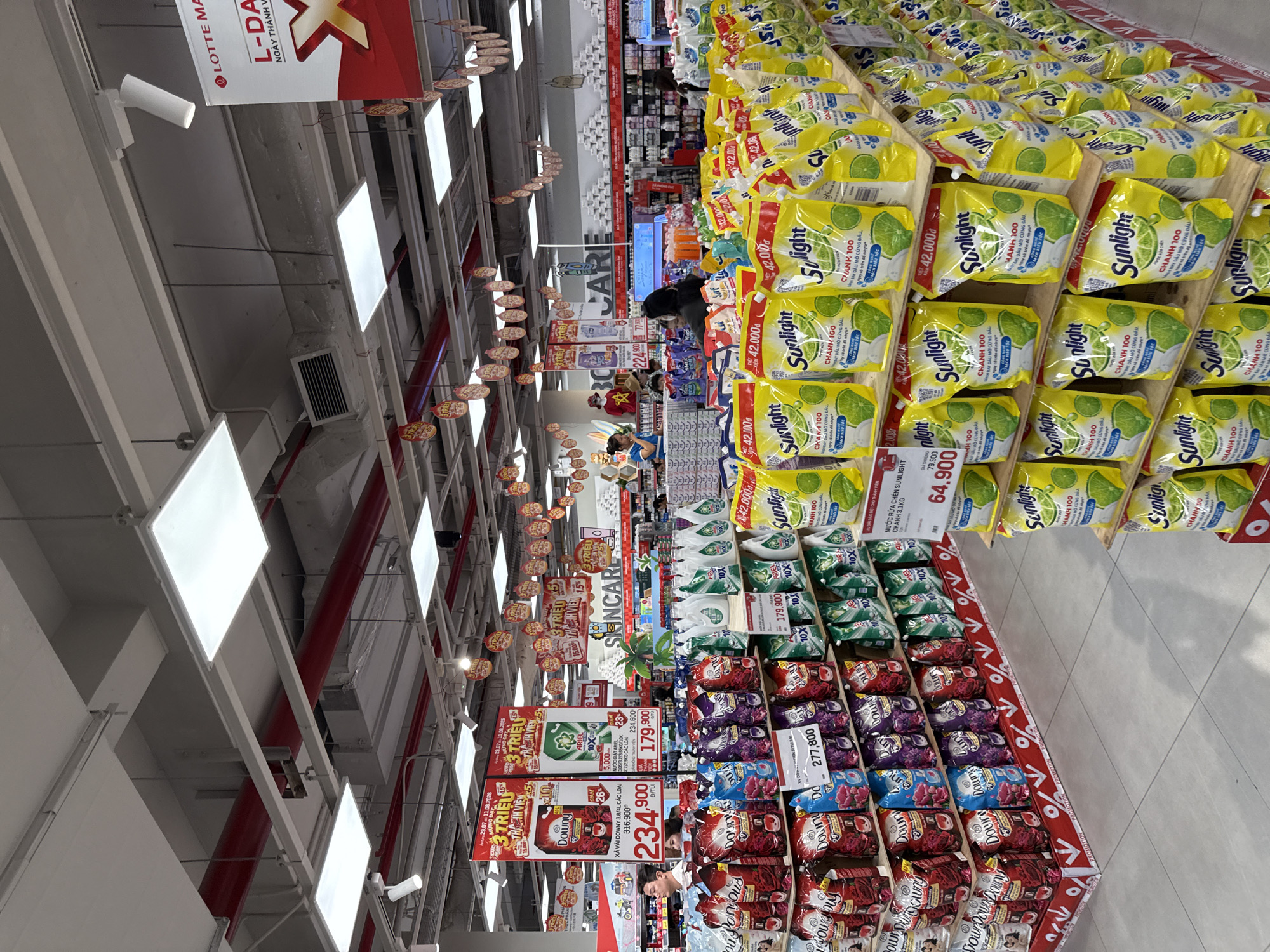 The image depicts the interior of a large supermarket, likely a Lotte Mart, given the signage at the top right. The store is well-lit with fluorescent ceiling lights and track lighting. The aisles are stocked with a variety of consumer goods, primarily laundry detergent and fabric softener.
In the foreground, large displays of yellow-packaged "Sunlight" brand laundry detergent pouches are prominently featured. These pouches are arranged in multiple tiers on wooden shelves. The price "42,000đ" is visible on several of these packages. To the left of the Sunlight detergent, there are shelves filled with red, blue, and purple-packaged fabric softener, labeled "Downy." Prices such as "234,900đ" and "179,900đ" are displayed for these products. Further left, more laundry detergent products are visible, including green-packaged "Ariel" detergent, priced at "179,900đ."
In the mid-ground, several shoppers are present, some looking at products and others walking through the aisles. A store employee wearing a blue shirt and a name tag appears to be interacting with a customer near a display of products. In the background, more shelves stocked with various household items are visible, extending deep into the store. A prominent banner with red and white text, including the "LOTTE MART" logo, is hanging from the ceiling. Other promotional signage and hanging decorations are also visible throughout the store. The floor is tiled in a light grey pattern. The overall environment suggests a busy retail setting. Based on the context of Lotte Mart locations in Vietnam and typical product offerings, this scene is likely set in Nha Trang, Vietnam.