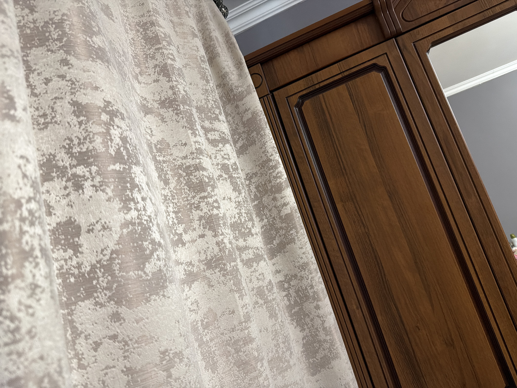 The image shows a close-up view of a textured, light-colored curtain on the left and a dark wood cabinet with ornate trim on the right. The curtain has a mottled pattern in shades of beige and white. The cabinet appears to be made of polished wood with raised paneling and decorative molding. The scene is indoors, with a hint of a ceiling and a gray wall in the background.