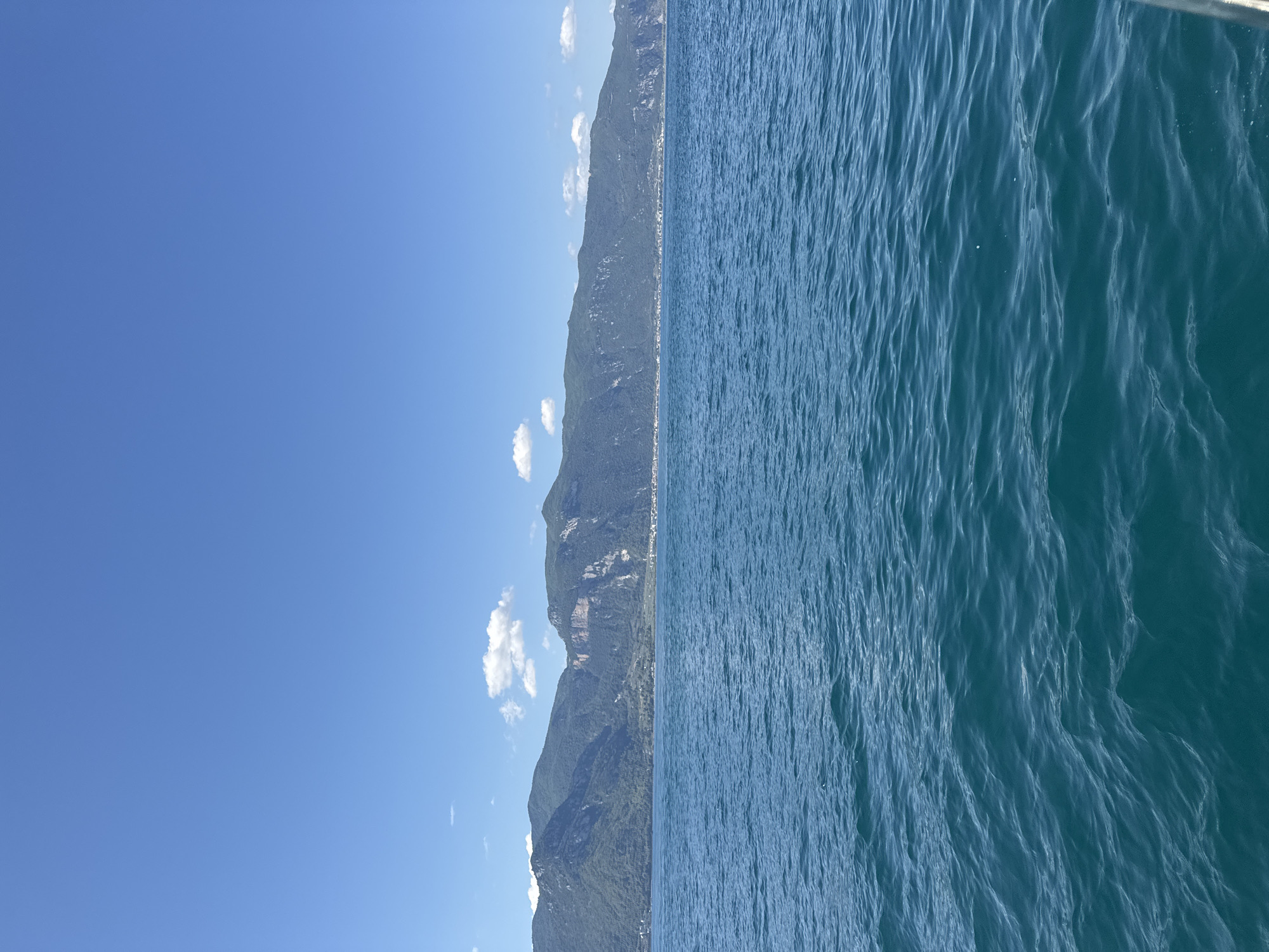 A wide shot captures a bright, sunny day with a clear blue sky above a tranquil body of water and a lush green mountain range. The water, a deep turquoise hue, shows gentle ripples across its surface. In the distance, a long, undulating mountain range dominates the horizon, its slopes covered in dense green vegetation. A thin line of buildings is visible along the base of the mountains, suggesting a settlement. A few scattered white clouds drift lazily in the sky. The scene appears to be a scenic landscape, likely a lake or the sea, with mountains forming a natural backdrop. The image is an exterior shot, taken during the daytime, with no people or specific actions depicted. The overall impression is one of peaceful natural beauty.