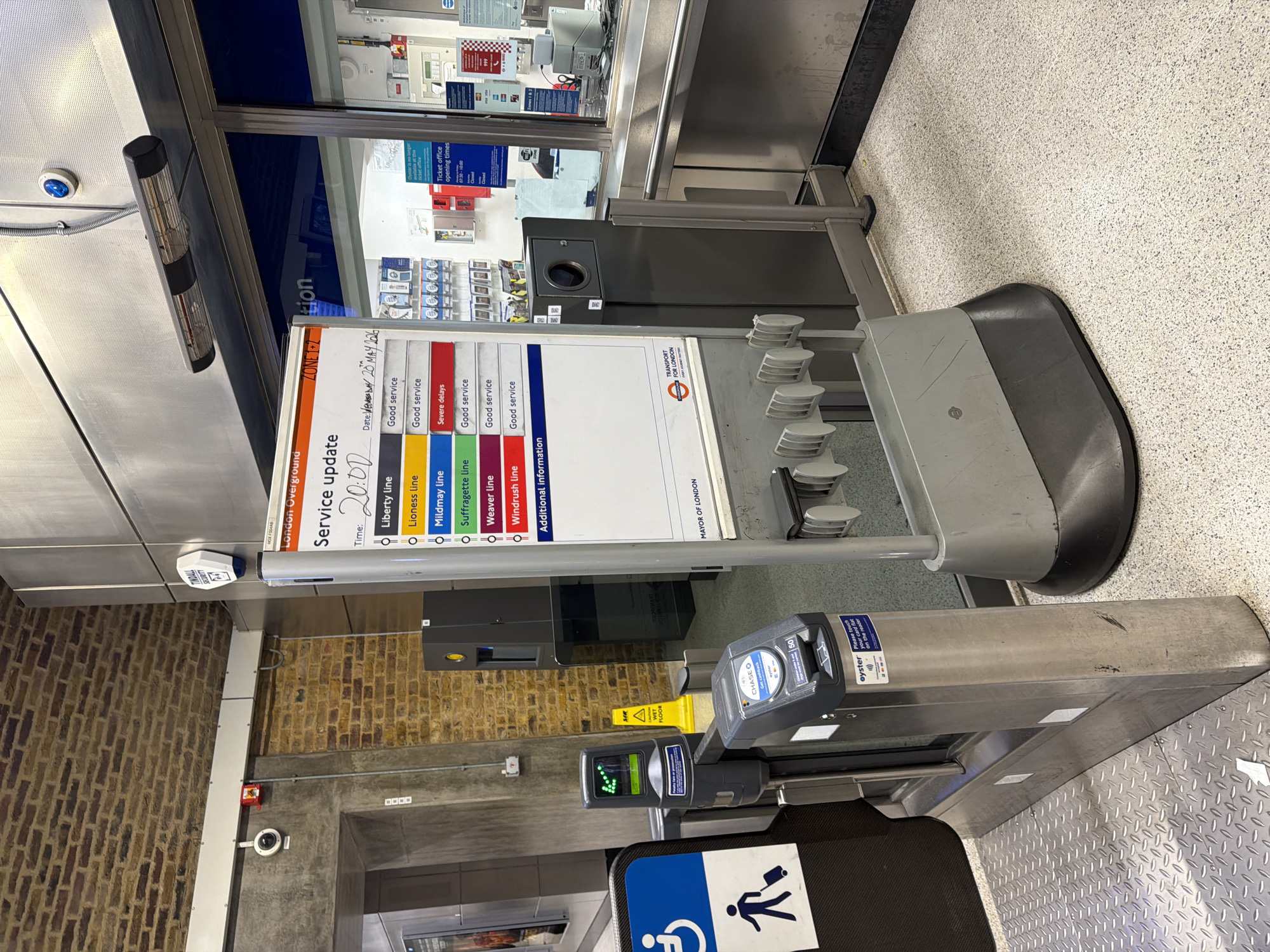 The image captures a scene inside a London Overground station, specifically at a ticket barrier. A service update display indicates that the date is Wednesday, May 20, 2026, and the time is 20:00 (8 PM). The display reports the service status of several lines: Liberty and Lioness lines are marked as "Good service," Mildmay line has "Severe delays," and Suffragette, Weaver, and Windrush lines are marked as "Good service." The display is near a ticket office with listed opening times and a sign indicating "Zone 1+2."
Adjacent to the display is a ticket barrier with an Oyster card reader. A wheelchair accessibility sign is visible. The background includes a brick wall and tiled flooring. The general environment suggests an urban transport hub within London.