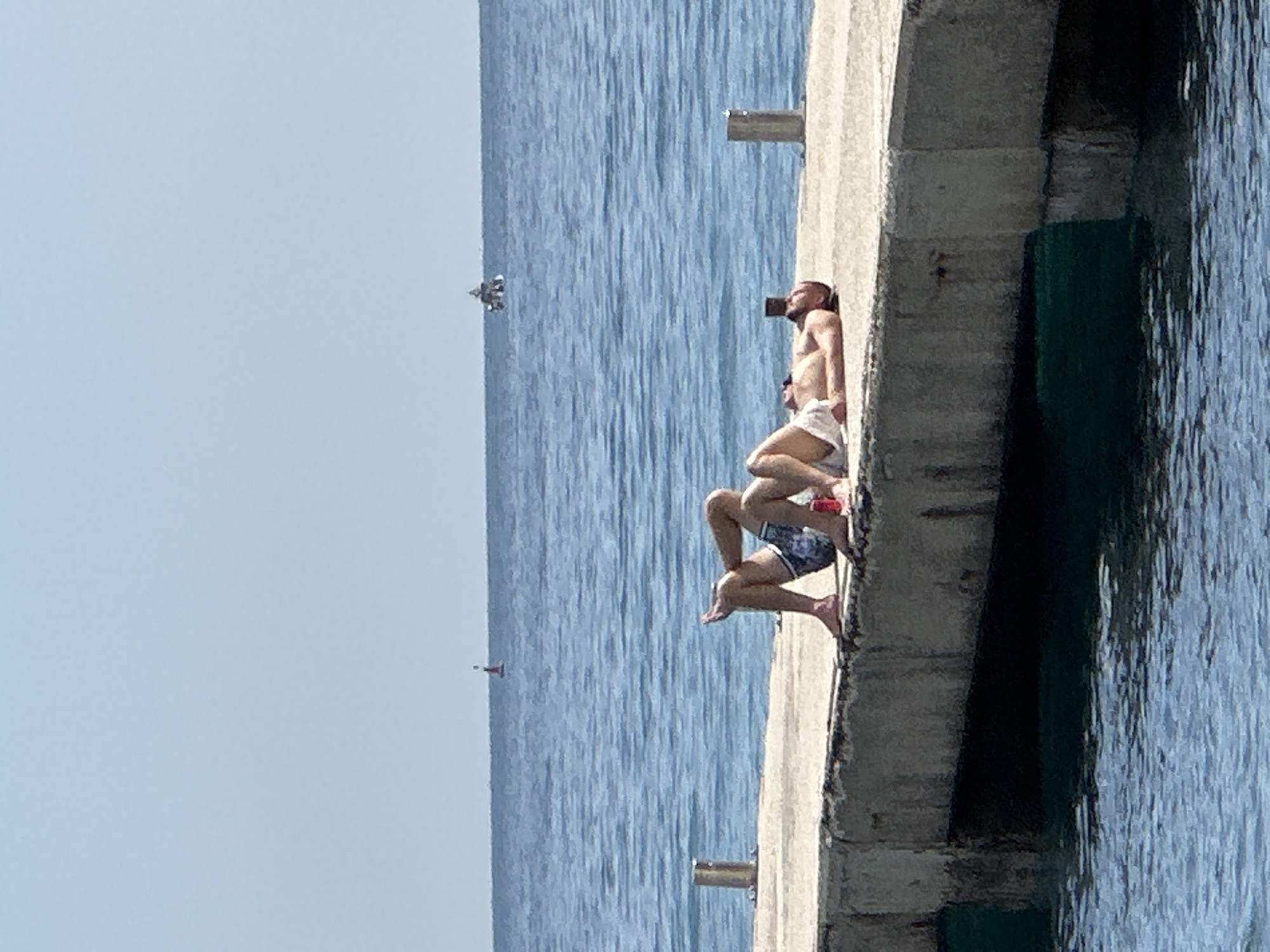 Two people are sunbathing on a concrete pier. The man is lying back, and the woman is leaning against him. In the sea, in the distance, is a small boat. In the sky, an airplane is visible. The location is Budva, Montenegro.