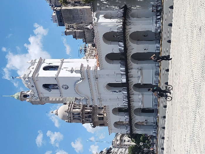 An outdoor scene in Buenos Aires, Argentina, depicts the historic El Cabildo on Plaza de Mayo under a clear blue sky with scattered white clouds. The white, colonial-style Cabildo building occupies the mid-ground, featuring a central clock tower with two visible clock faces (one indicating approximately 9:00) and a series of arched ground-floor openings leading to a prominent upper-level balcony with a black iron railing. Behind and to the left of the Cabildo's tower, the Palacio de la Legislatura de la Ciudad de Buenos Aires is visible with its light-colored dome and classical architecture. To the right, other multi-story urban buildings rise. The immediate foreground consists of a large, textured paved plaza. Two adult males are present in the foreground: one is seated on and operating a dark folding bicycle, positioned towards the right; the other stands to the right of the bicyclist, facing away from the camera, with his right hand raised towards his head. A line of dark conical bollards separates the plaza from a road marked with white lines directly in front of the Cabildo. Additional indistinct figures are visible on a street to the far left.