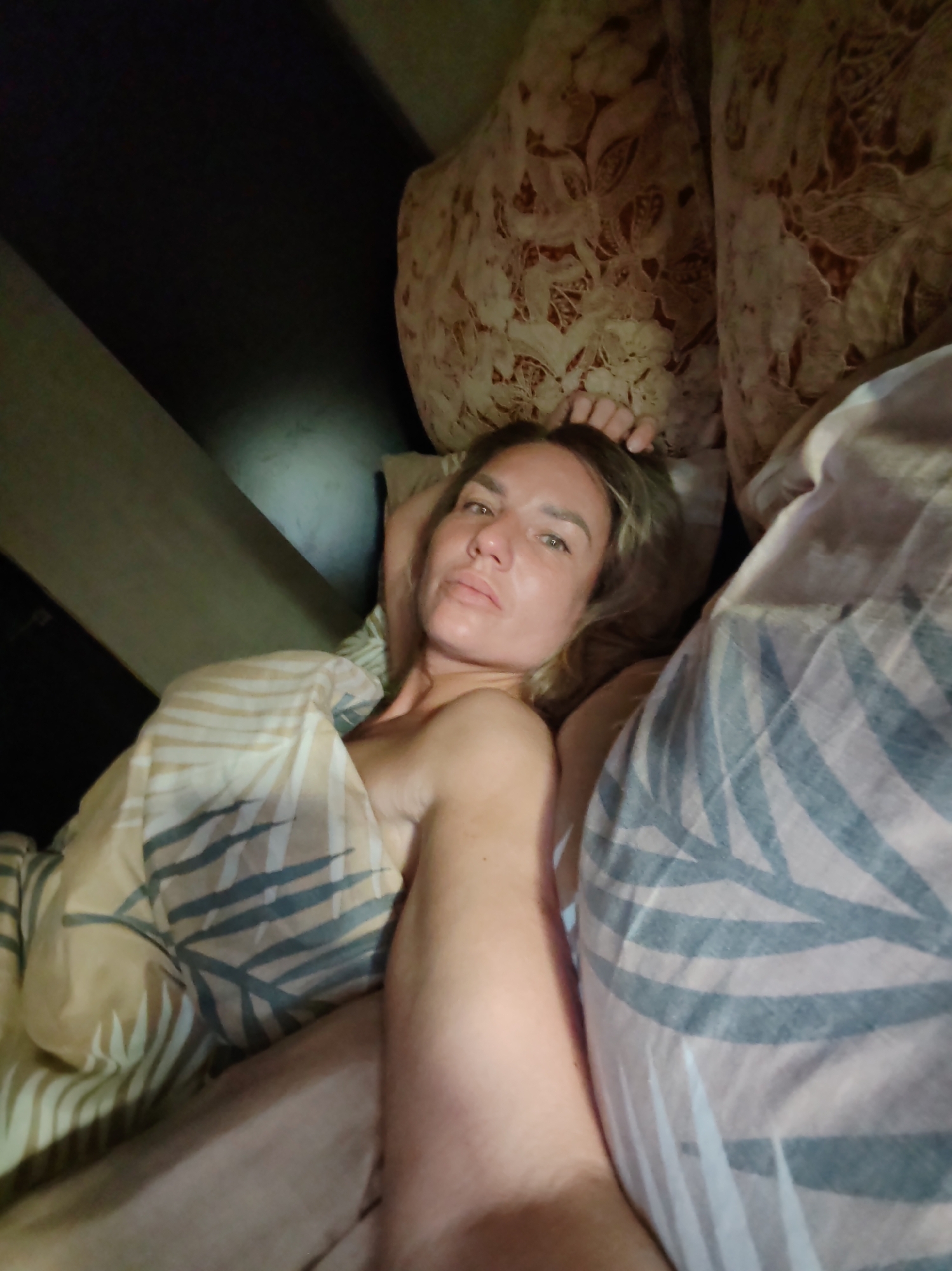 A woman with blonde hair and fair skin is lying in a bed, partially covered by a patterned blanket. She appears to be looking directly at the camera. The bed is surrounded by pillows with a floral pattern, and there is a dark, indistinct area in the background, possibly a wall or furniture. There is no information available to determine the location is in Petropavlovsk-Kamchatsky, Russia.