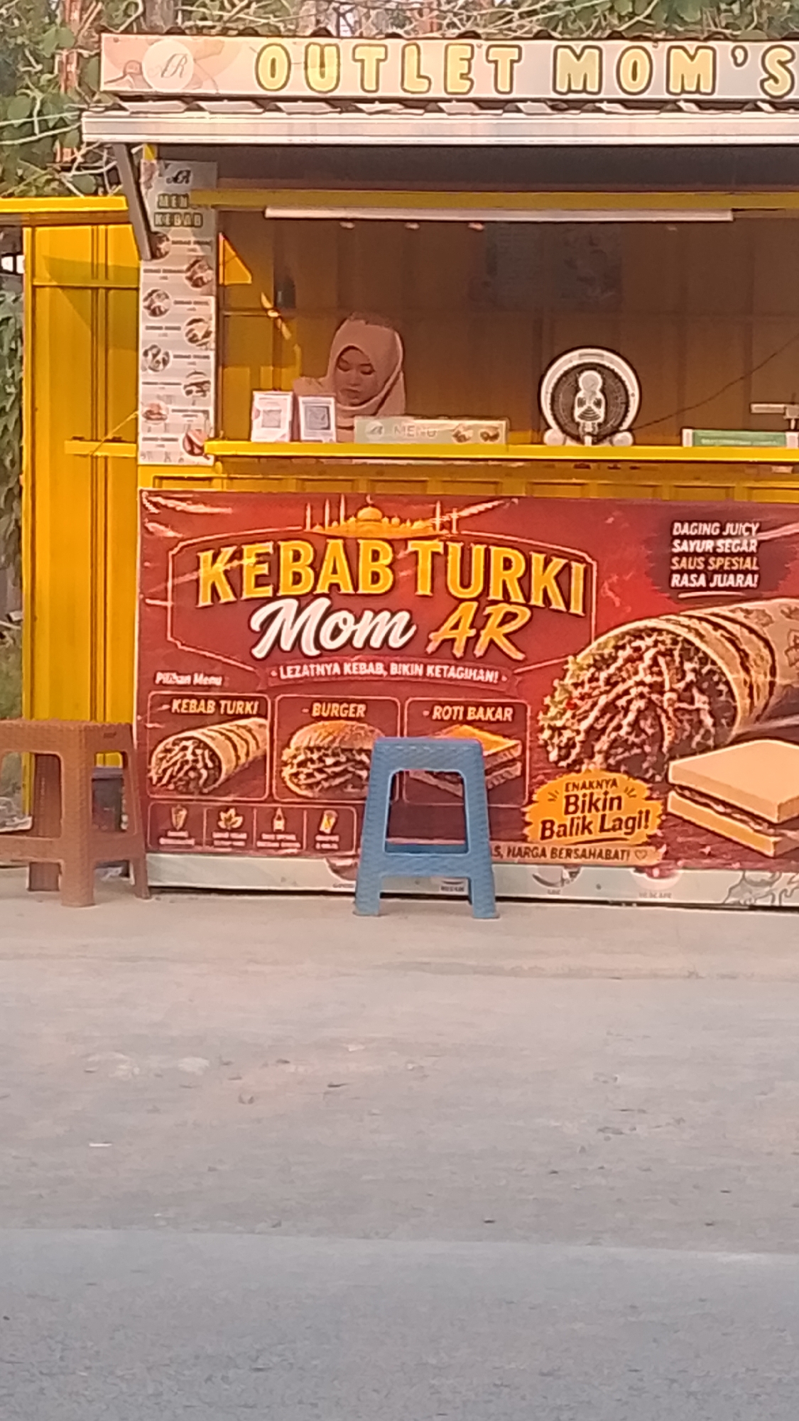 The image shows a food stall in Karanglangu, Indonesia, that sells "Kebab Turki Mom AR". The stall is yellow and has a banner with pictures of kebabs, burgers, and roti bakar. A woman wearing a hijab is visible behind the counter. Two small stools are placed in front of the stall. The stall is located on a street with a paved surface. The background shows trees and buildings.
