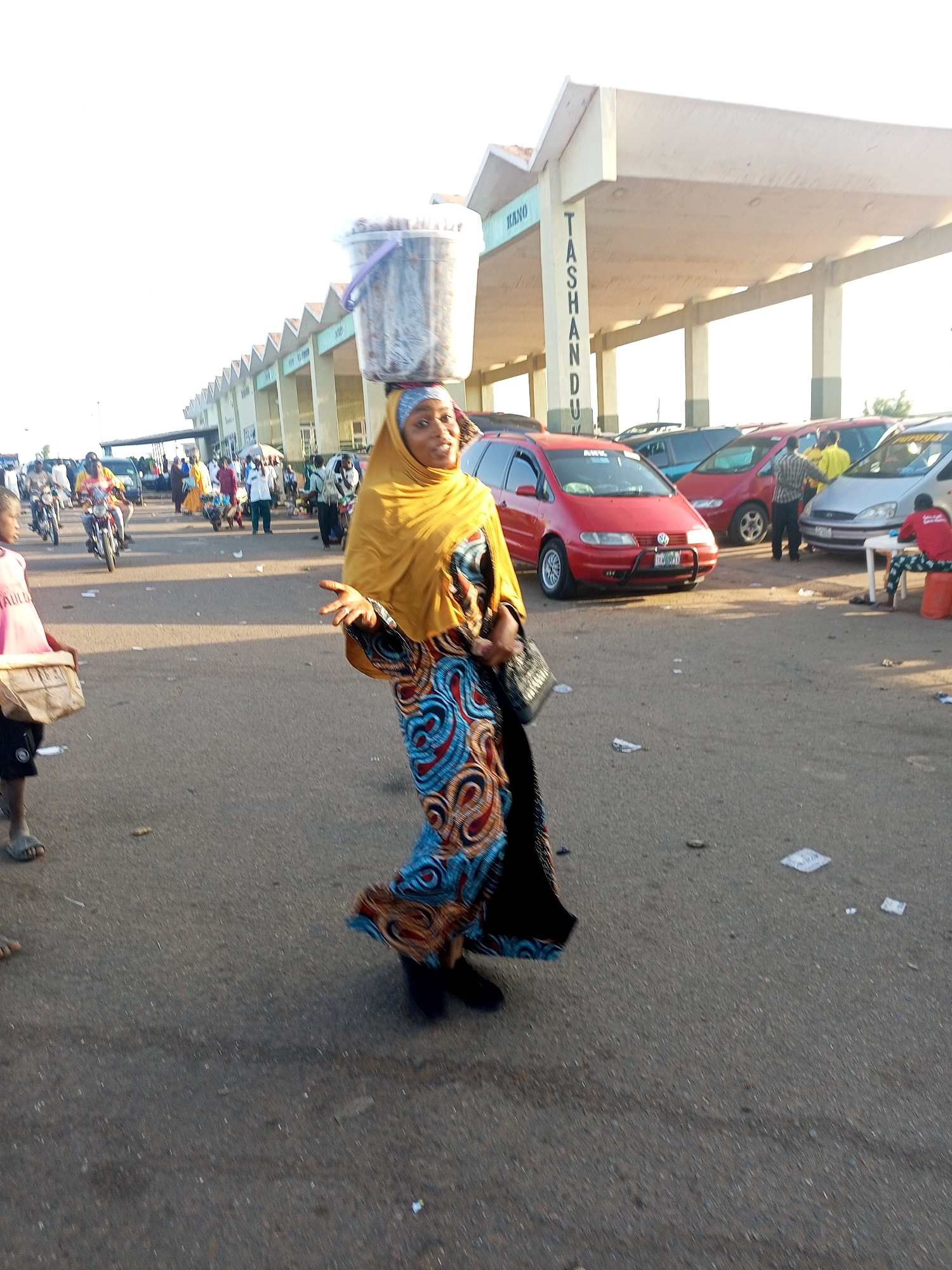 In a city identified as "KANO TASHANDU" on a structure, a woman carries a bucket on her head. She is wearing a yellow headscarf and a patterned blue, orange, and black dress. She is positioned on a paved area littered with debris. To her left, a child is partially visible. Behind her, there are people, parked cars, and motorbikes.