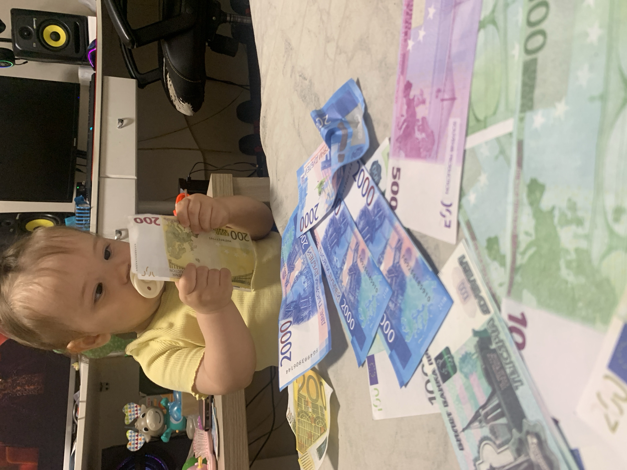 A baby wearing a yellow t-shirt sits at a table and holds a 200 Euro banknote to their mouth. In front of the baby are several Russian Ruble banknotes, including 2000 Ruble notes, and Euro banknotes of 100, 500, and green notes, possibly 100 Euro notes. The background shows a desk with a computer monitor and speakers, and a black office chair. The scene appears to be indoors, likely a home environment. The location of Chaikovsky, Russia, is not explicitly visible or inferable from the image itself.