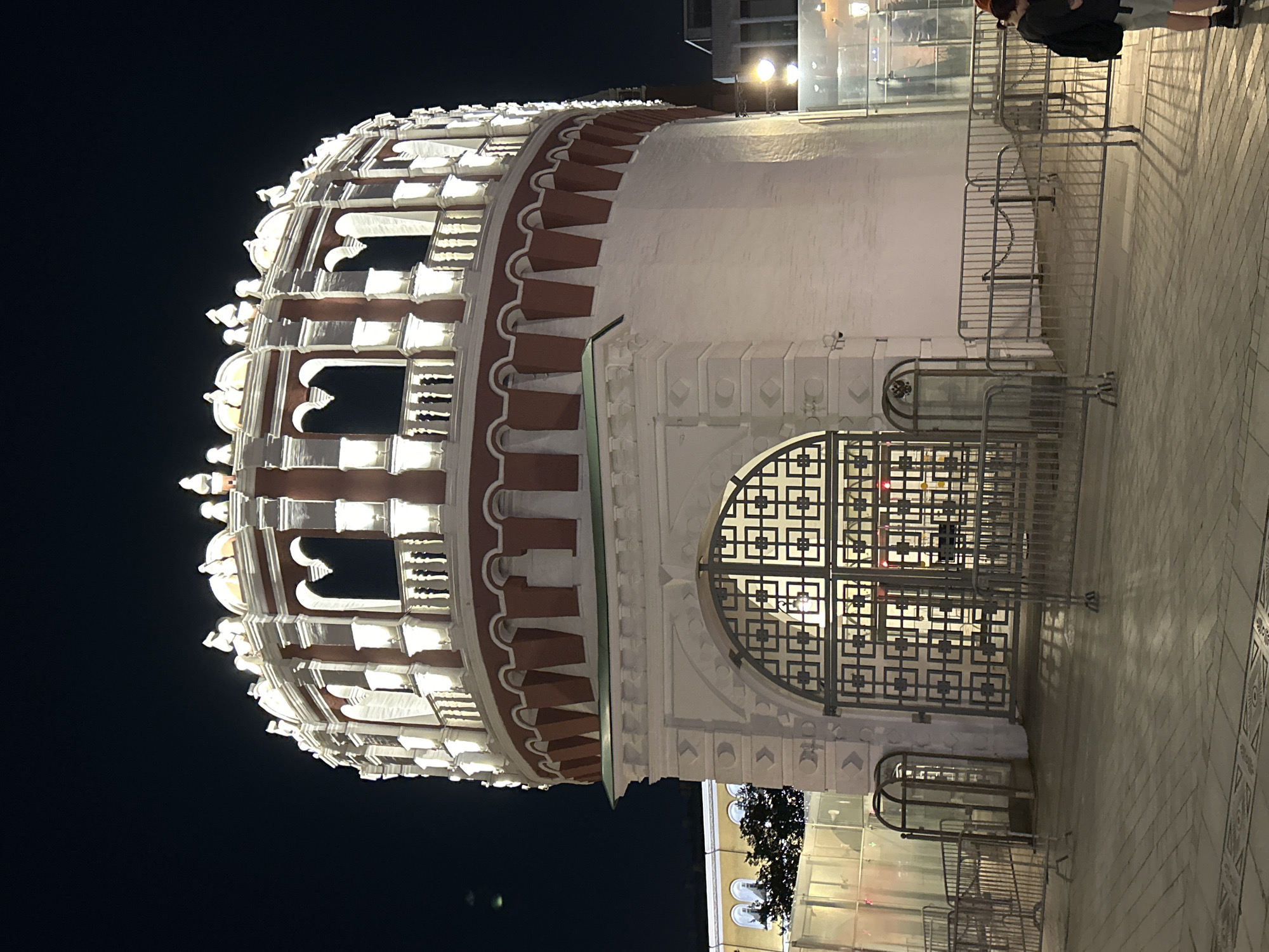 An exterior night shot captures a large, circular, white brick tower with red trim and white decorative arches at its upper level. The tower is illuminated by numerous lights. In front of the tower is a large, ornate, dark metal gate. The foreground consists of a paved walkway and metal barricades. To the left of the tower, a yellow building with arched windows is partially visible. Two people are standing on the right side of the walkway, near the barricades. The sky above is dark.