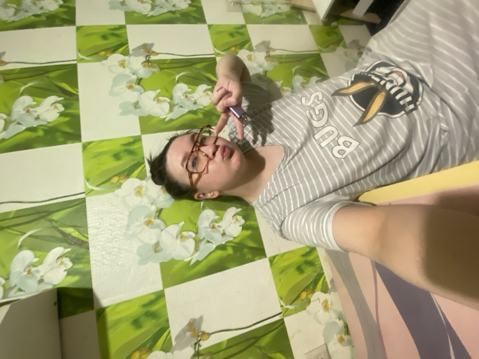 The image depicts an indoor scene in what is identified as the city of Chaikovsky, Russia. A single individual, presumed female, with light skin and dark hair styled in a bun, occupies the central frame. She wears square-framed eyeglasses with amber-colored rims and a grey and white striped short-sleeved garment, possibly a dress or long t-shirt, featuring the word "BUGS" and a graphic of a cartoon rabbit wearing sunglasses on the chest. The individual holds a small, light purple, rectangular object in her right hand, simultaneously extending her index and middle fingers in a V-sign. Her lips are pursed in a pouting expression directed towards the viewer. She appears to be seated or leaning, with a yellow surface visible to her right and a pink and purple striped surface in the immediate foreground. The background consists of a wall covered in wallpaper patterned with alternating green and white squares, where the green squares display images of white orchid flowers with green stems and leaves. A white electrical outlet or switch is partially visible on the far left wall, and a segment of white furniture is present on the far right.