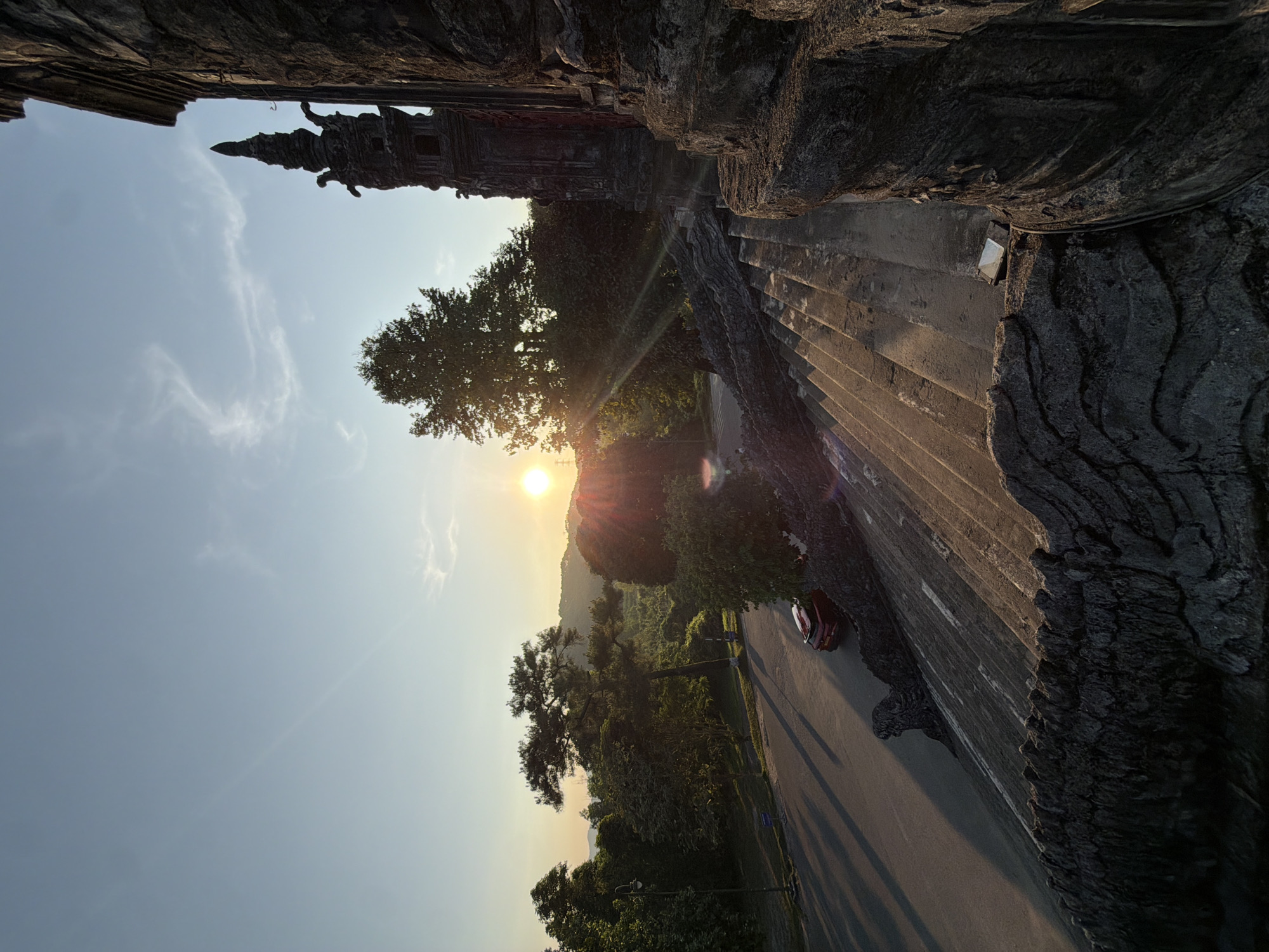 The image depicts a view in Thủy Xuân Ward, Vietnam, captured at dusk or dawn. A multi-tiered tower stands silhouetted against the sky in the upper left. The sun, appearing low on the horizon, is partially obscured by a tree, casting rays across the scene. A road runs through the frame, with a single car visible. The right side of the image is dominated by a textured, wall-like structure, possibly part of a building or monument. The foreground features dense foliage.