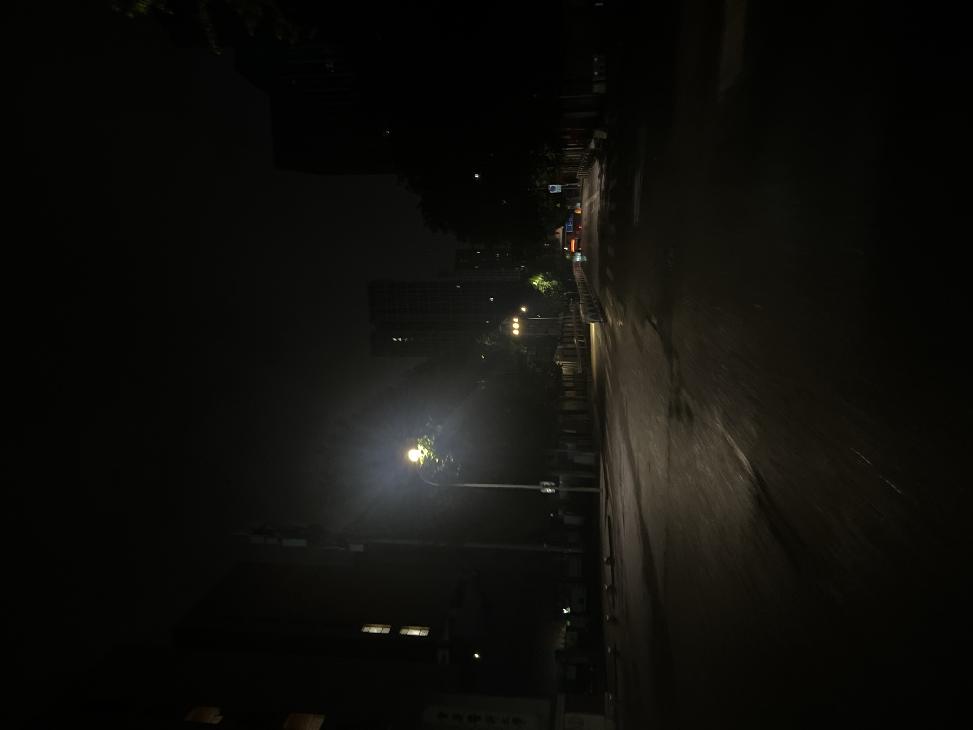 This image captures a nighttime street scene in Yuzhong District, Chongqing, China. The asphalt road, appearing wet and reflective, recedes into the distance under dark conditions. A prominent street lamp on the left side of the mid-ground emits a bright, warm yellowish-white light, illuminating a portion of the road and the foliage of a nearby tree.
On the left, a multi-story building is visible, featuring several rectangular windows with interior lights on. Partially visible Chinese characters on a vertical sign on this building read "重庆医科大学" (Chongqing Medical University). A utility pole or similar structure stands between this building and the road. On the right side, a line of trees borders the road, with taller, darker buildings in the background displaying scattered illuminated windows.
Further down the road, distinct red taillights indicate the presence of a vehicle moving away from the viewer. Blue and white road signs are visible on the right, alongside orange and white temporary barriers or cones, suggesting a roadwork area or temporary lane alteration. Several low, light-colored bollards or barriers are positioned along the left edge of the road in the foreground. No individuals are present in the depicted scene.