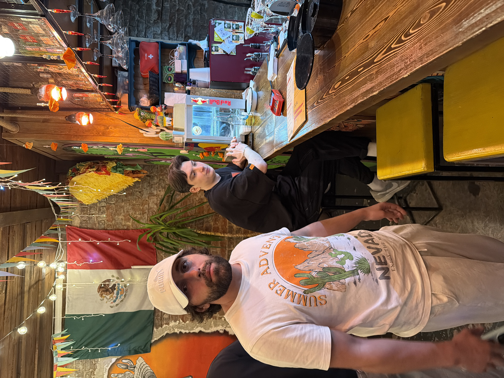 Two males are visible in what appears to be a bar or restaurant. One is seated at a wooden bar with yellow bar stools; he is wearing a black shirt and dark pants. The other male is standing closer to the camera and wearing a white t-shirt with desert themed text and illustrations along with beige pants, and a white Puma visor. A Mexican flag hangs on the wall behind him, adorned with string lights and colorful pennant banners. Additional decorations and bar equipment are visible in the background. The location appears to be "Mama's", a restaurant in Krasnoyarsk, Russia.