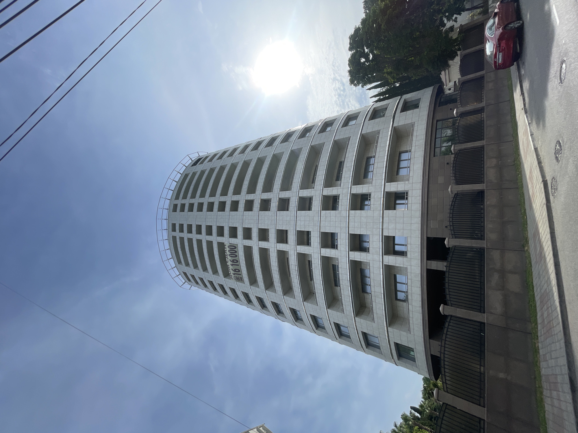 An eye-level, wide-angle, upward shot captures a modern, circular, light-colored apartment building on a sunny day in Sochi, Russia. The building, with numerous balconies, dominates the frame, reaching towards a clear blue sky with sparse clouds. A bright sun is visible in the upper right corner. A black metal fence encloses the property, with a red car parked on the street in front of it. The street is paved, with a sidewalk and some greenery, including cypress trees, visible. A sign on the building reads "ДОМ СДАН 81965 16 16 000", indicating that the building is completed and providing a phone number. Overhead power lines are visible in the upper portion of the frame.
