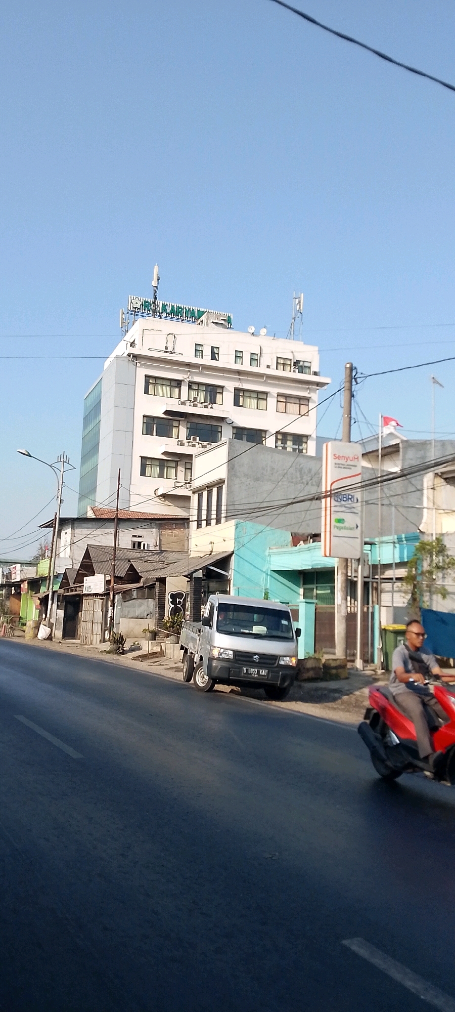 The image captures a street scene in Bekasi, Indonesia, likely during daylight hours given the clear blue sky and shadows. Dominating the upper portion of the image is a multi-story white building with a green sign at its peak that reads "RAG KARIMUN." The building has numerous windows and appears to be an office building or hotel. In front of it, a smaller, older structure with a corrugated metal roof is visible, possibly a row of shops or houses.
To the right of the older structure, a silver Suzuki pickup truck is parked on the side of the road, facing away from the camera. Its license plate is visible and reads "D 9053 KAU". Further to the right, a street sign with "Senyum" and "BRI" is mounted on a pole, indicating a bank branch. A red and white Indonesian flag is visible flying behind the sign.
In the foreground, a person is riding a red motorcycle, moving from right to left across the frame. The rider, wearing glasses and a grey shirt, is partially visible. The road itself is asphalt with a white line marking the lane. The overall impression is of a moderately busy urban environment with a mix of modern and older architecture.