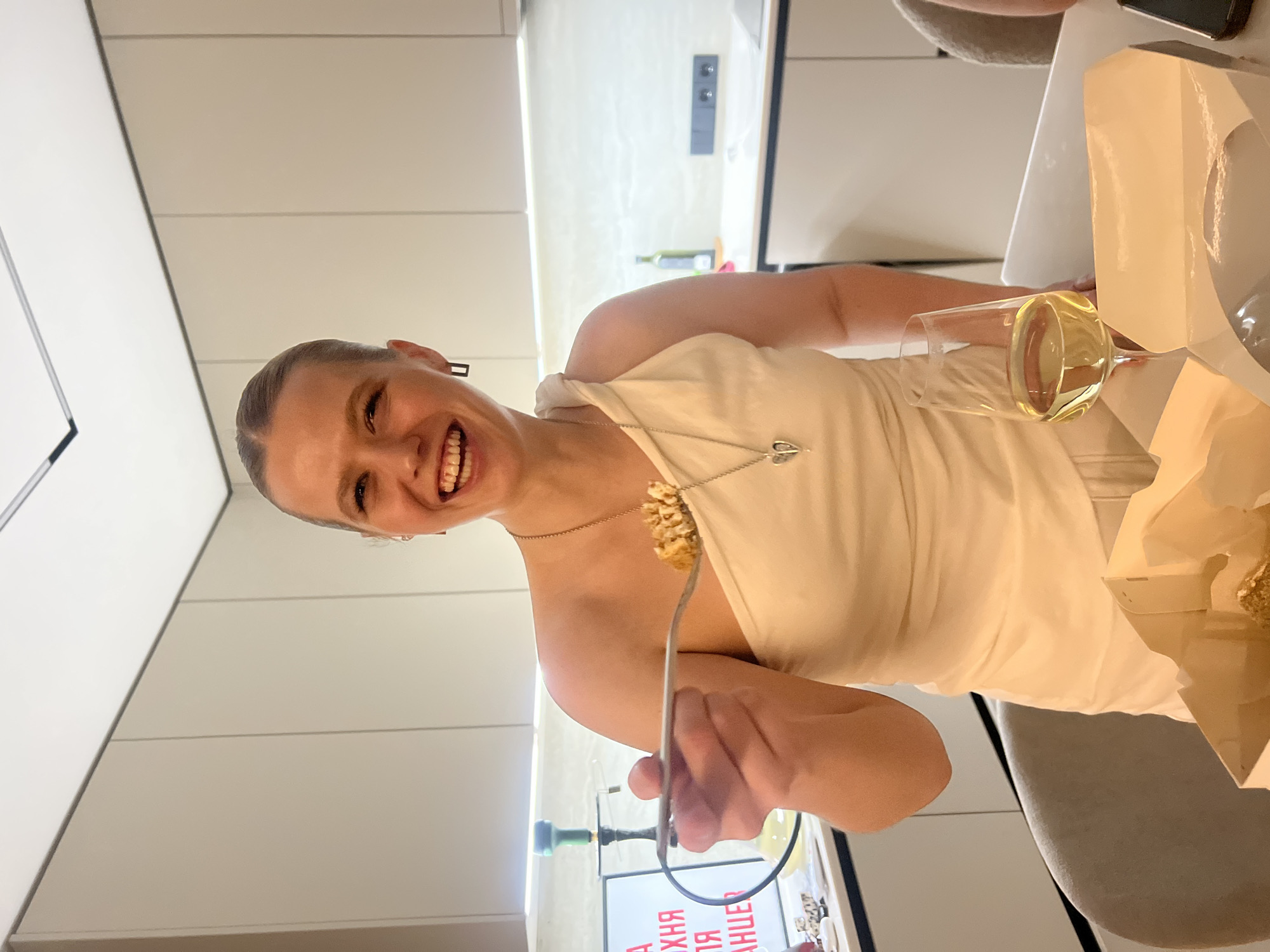 A woman is indoors, facing the camera with a smile. She is wearing a light-colored dress and a necklace. She is holding a fork with food on it. A glass of white wine is on the table to her right. In the background is a modern kitchen with light-colored cabinets. Text in Cyrillic is visible on an electronic device in the background, indicating a possible location in Russia. The city mentioned, Рубцово, Russia, cannot be verified as the actual location based on the image alone.