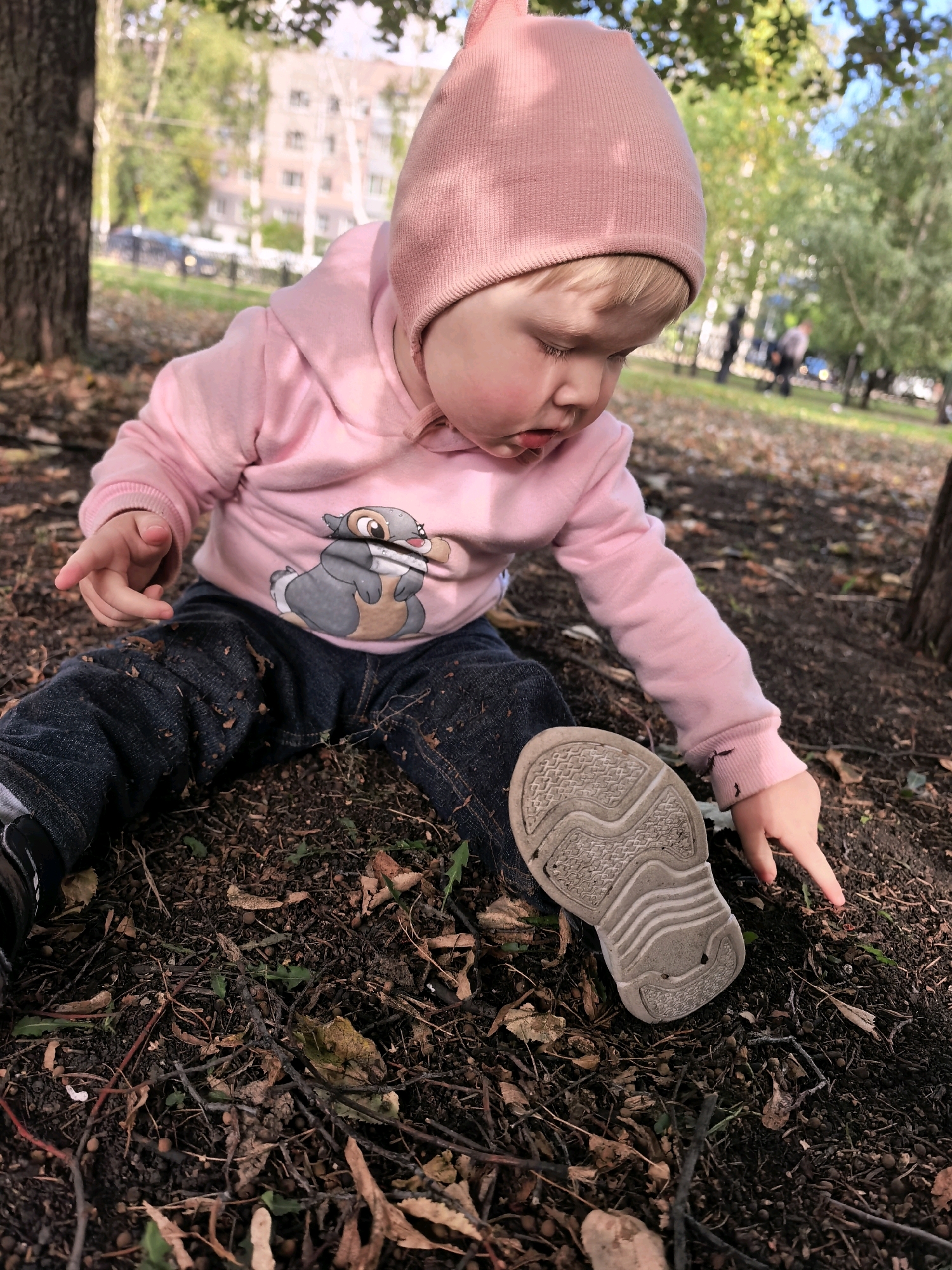 A young child wearing a pink hooded sweatshirt and jeans sits on the ground outdoors. The child is looking down at the ground, with their right hand extended towards the soil. A white sneaker is visible on the child's right foot. The ground is covered in dirt, fallen leaves, and small twigs. In the background, trees and buildings are visible, suggesting an urban park setting. The lighting indicates it is daytime.