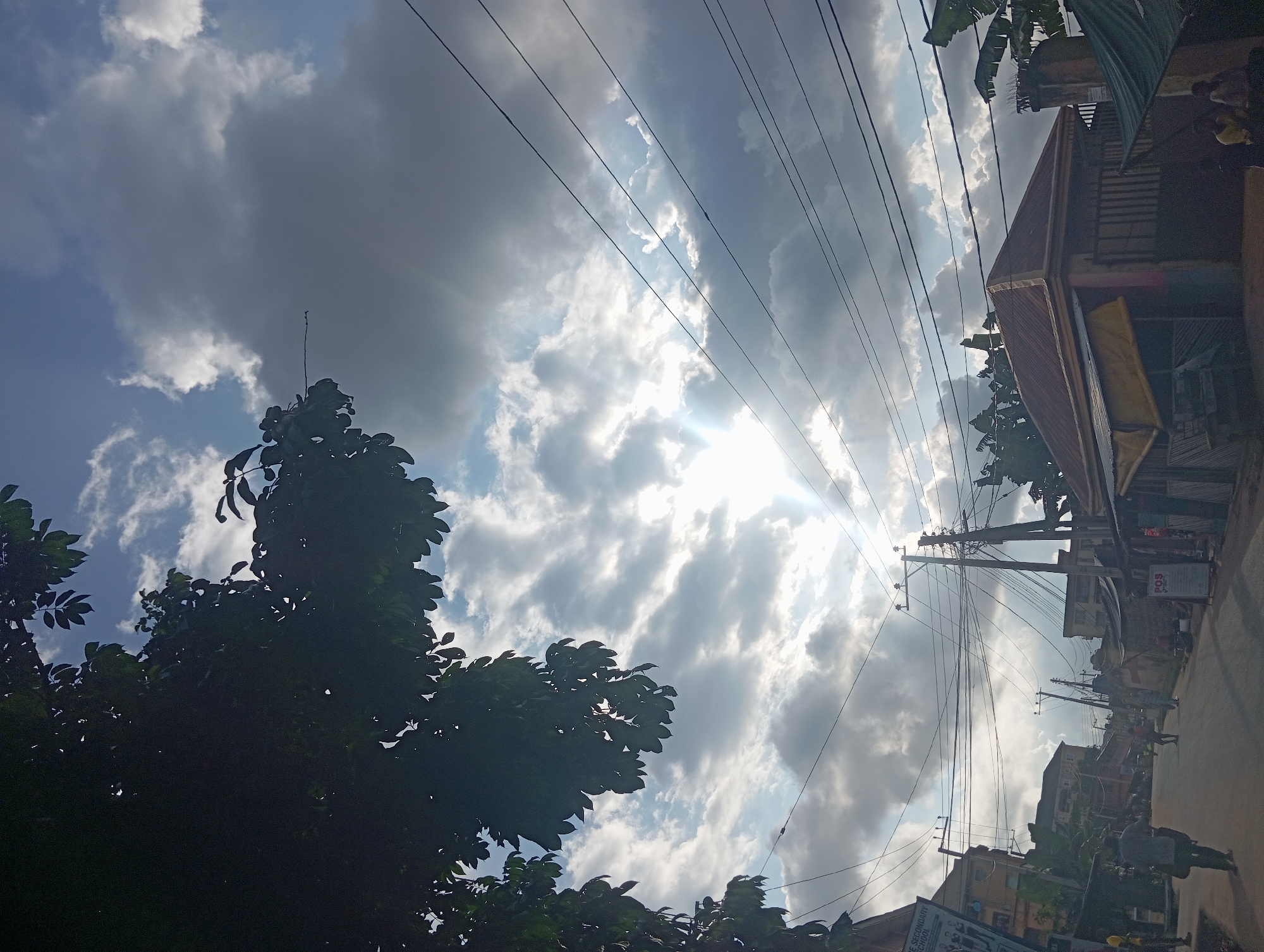 The image shows a street scene in Nkpor, Nigeria, under a cloudy sky. The sun is partially obscured by clouds, casting rays of light. A large tree with dark green leaves is visible in the foreground on the left, partially shading the view. Power lines are strung across the sky, supported by utility poles. Buildings line both sides of the street. On the right, a building with a reddish-brown corrugated metal roof has a yellow awning over a storefront. To the left of the street, there are other buildings, including a taller structure with visible windows. Several people are present on the street; one person in a grey shirt and dark pants walks away from the camera on the left side of the road. Further down the street, a few other individuals are walking or standing. The street is paved and appears to be a moderately busy urban area.