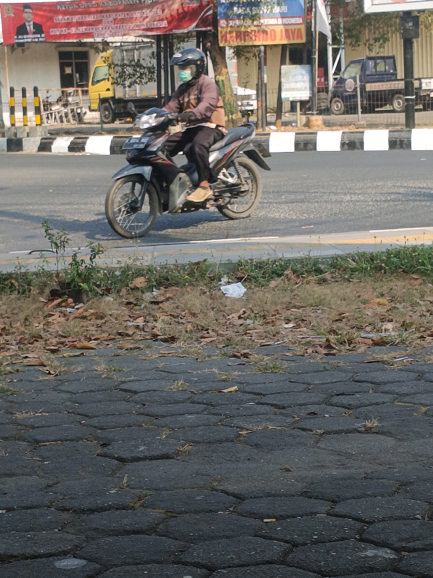 A person wearing a helmet and a face mask rides a motorcycle on a paved road. The person is wearing a brown jacket and dark pants, with tan shoes. The motorcycle is black and gray, with a license plate visible. In the background, a red banner with text and an image of a man is displayed on a building. A yellow truck and a dark blue pickup truck are parked behind the banner. The road has a striped white and black border. The foreground shows a sidewalk made of interlocking hexagonal pavers, with dry leaves and grass growing in between. The scene appears to be in an urban environment during daylight.