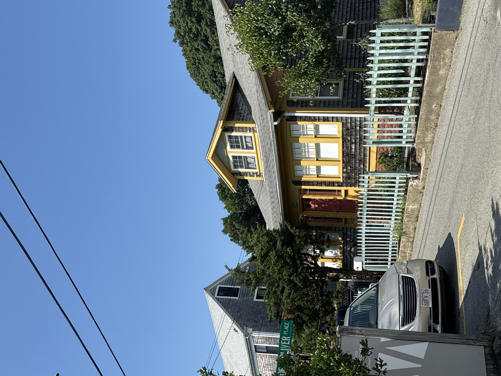 The image captures a sunny day in Provincetown, Massachusetts, United States of America. A silver minivan, bearing the license plate "5BFV 36", is parked on the right side of a paved road. The house directly behind the minivan is a single-story, grey-shingled residence with yellow trim around its windows and doors. A light blue, picket fence separates the house from the street. Further down the road, another house, also grey-shingled with a white roof, is visible. A green street sign indicating "OLIVER PLACE" is mounted on a post between the two houses. The sky is clear and blue, with power lines crisscrossing the upper portion of the frame. Trees and foliage surround the houses, suggesting a residential neighborhood.