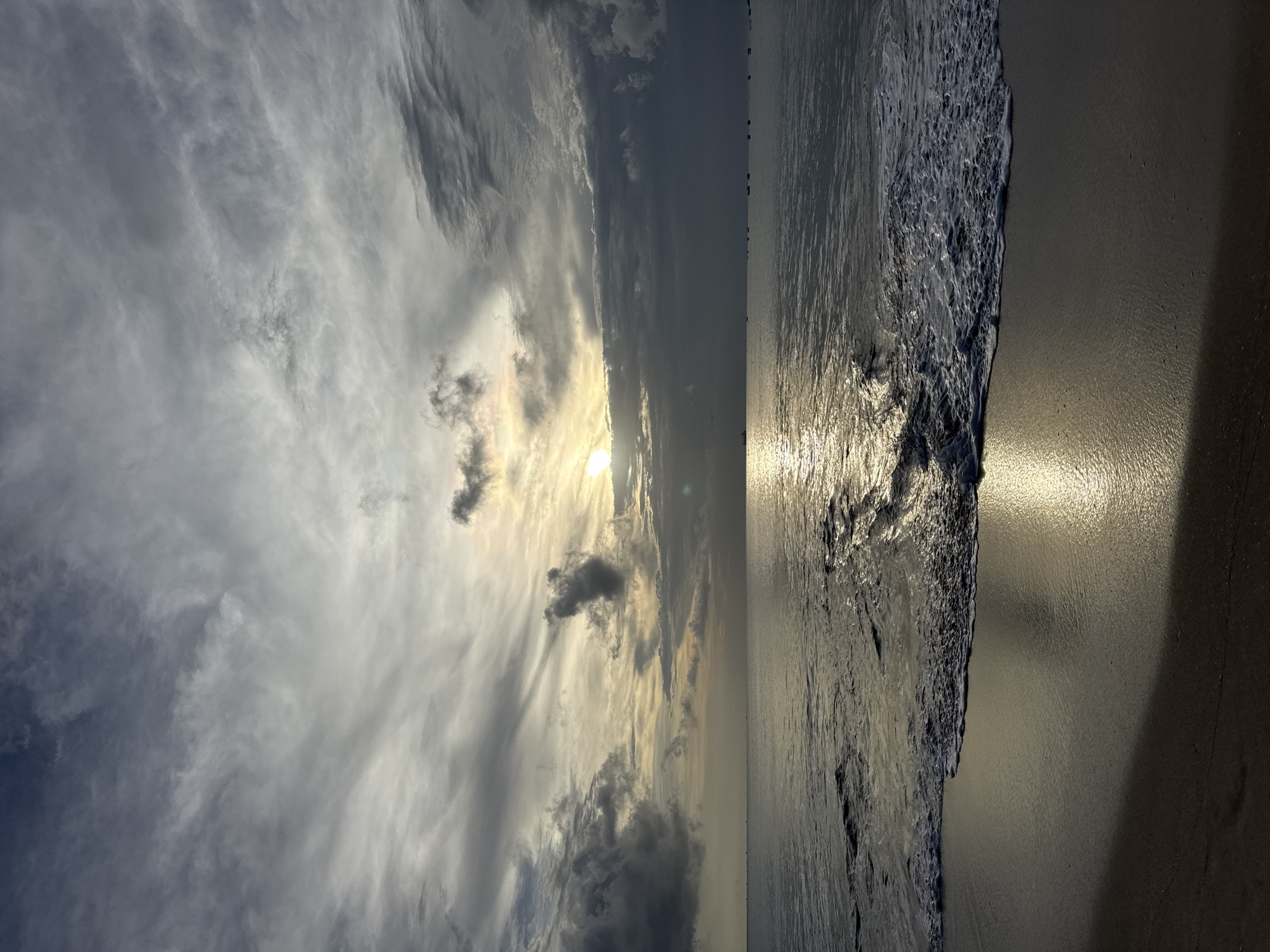 The image shows a beach scene, likely at sunset. The sky is filled with dramatic clouds, with the sun shining through them, creating a bright glow. The ocean reflects the sunlight, creating a shimmering effect. The beach is sandy, and the waves are gentle. The location is possibly Kuta, Indonesia, based on the context provided, although this cannot be confirmed from the image alone. There are no people visible in the image.