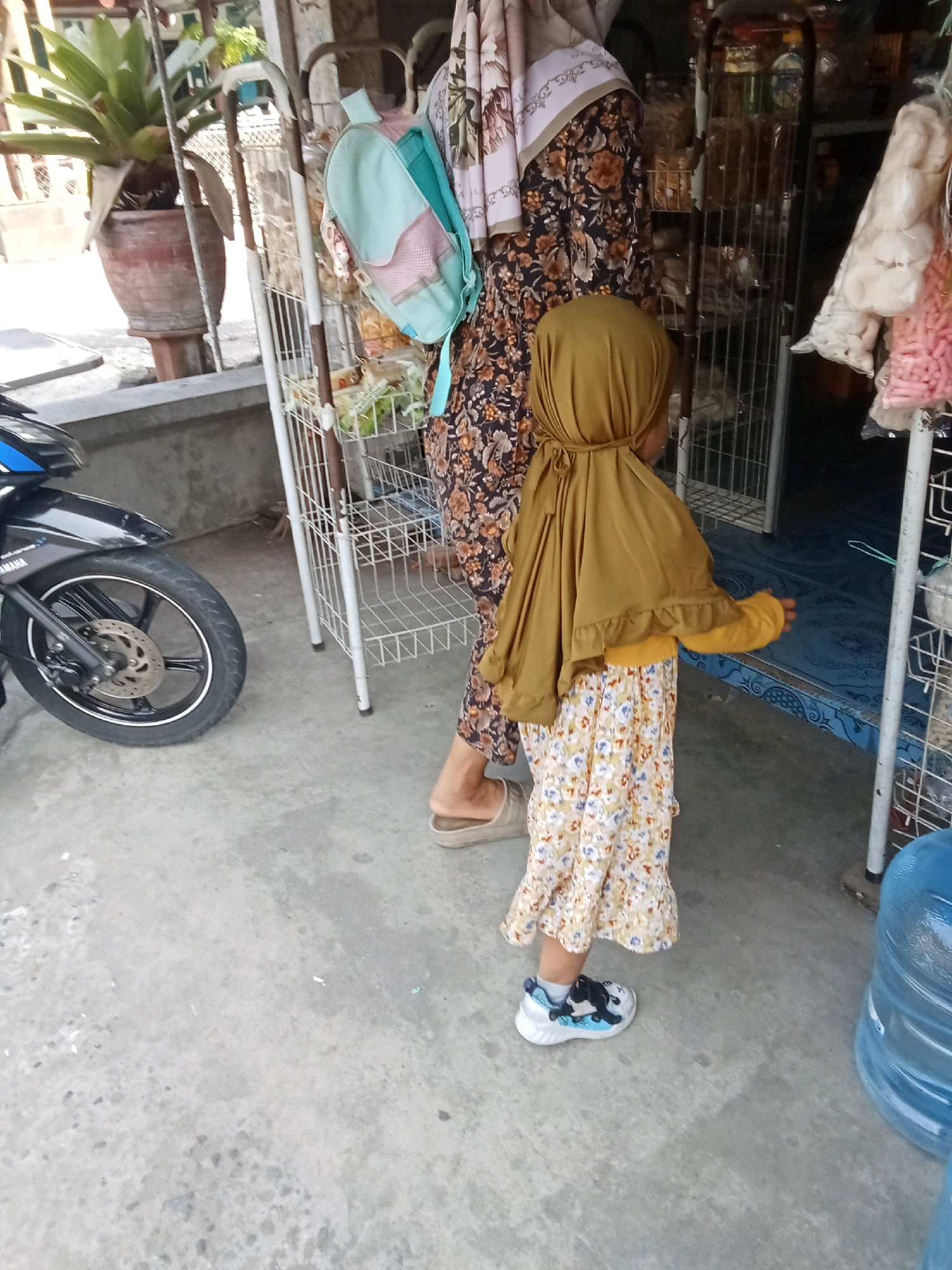 In Kaliwungu, Indonesia, a young child wearing a yellow hijab and a floral dress stands on a concrete ground. A woman, partially visible and wearing a patterned dress, stands beside the child. Behind them, a white metal shelf unit holds various items, and a turquoise backpack is attached to it. A black motorcycle is parked to the left of the scene, and a large blue water dispenser is visible on the right. The background includes a potted plant and the entrance to a shop.