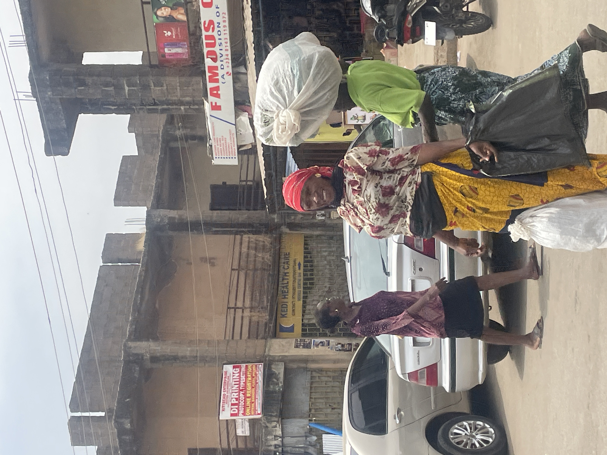 The image depicts a street scene in Jos, Nigeria. Two women are carrying large bundles on their heads and shoulders while walking along the sidewalk next to a parked silver car. The woman in front wears a red headscarf and a floral shirt with a yellow patterned skirt. The woman behind her wears a green shirt. In the background, buildings are visible with signs for "DI Printing", "Kedi Health Care", and "Famous (A Division of)". A motorcycle is parked nearby.