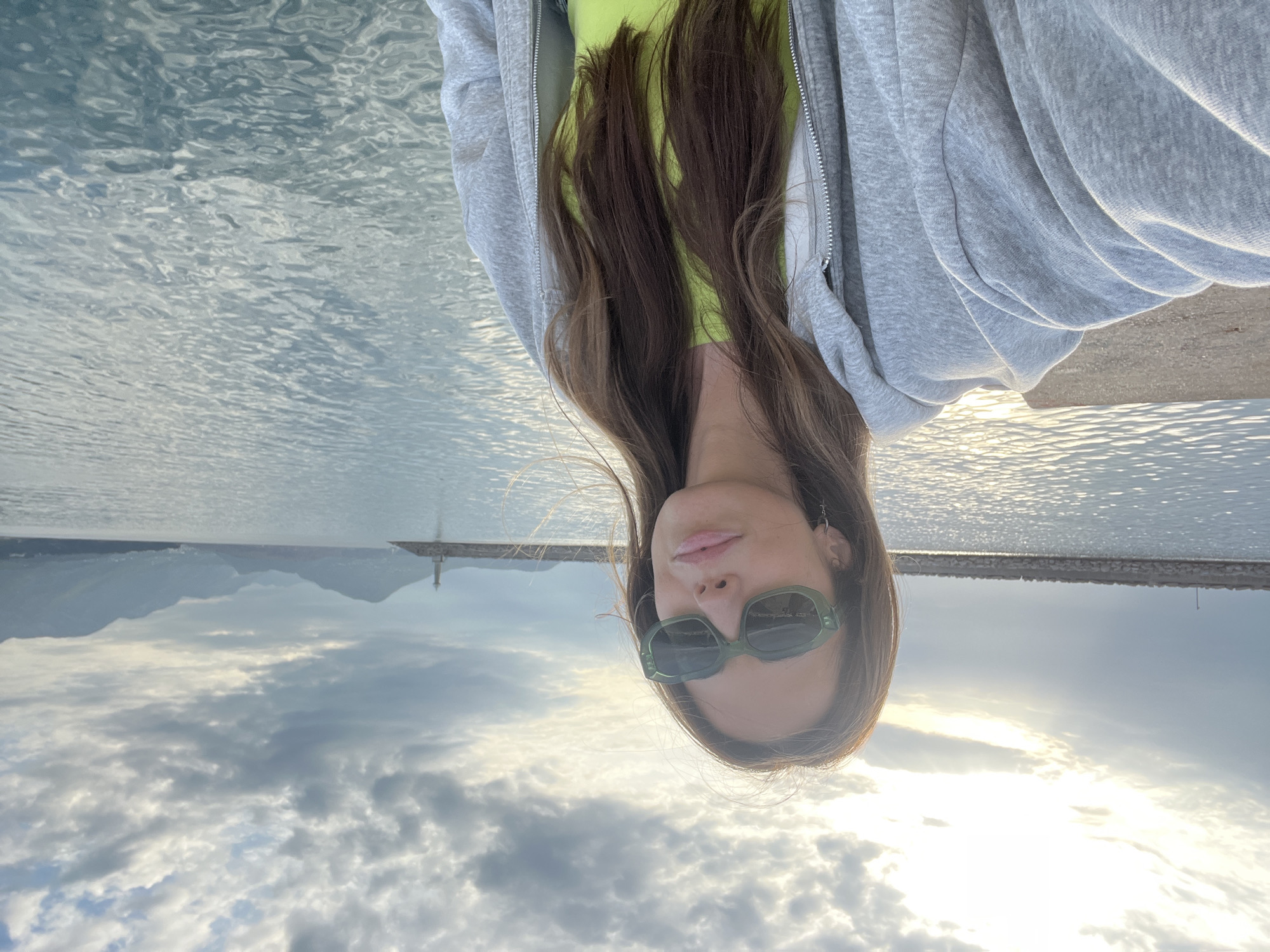 A woman wearing sunglasses, a lime green top, and a gray hoodie is facing the camera. Her brown hair is hanging down. Behind her, there's a view of the sea, with mountains and a lighthouse visible in the background. The sky is filled with clouds and the sun is shining through them. The location is likely in the city of Bar, Montenegro, given the presence of mountains, a lighthouse, and the sea.
