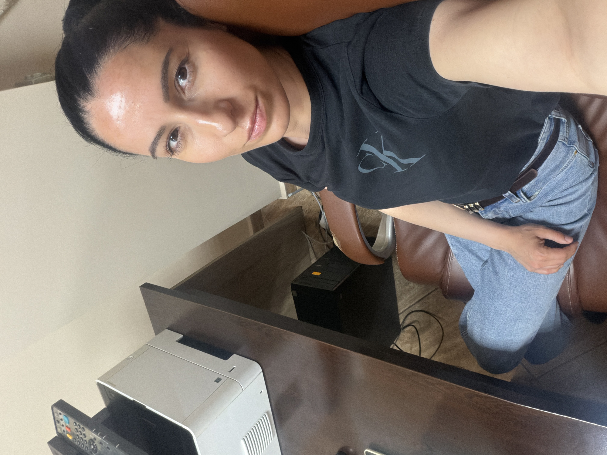 A woman with dark hair pulled back is seated in a brown leather chair. She wears a black T-shirt and light blue jeans. Her face is visible, with a slight smile. A dark brown desk with a printer on top is in the foreground. Behind the desk, to the right, is a black computer tower. The floor is tiled. This image was likely taken indoors, and there is no direct evidence to confirm the location as Pleven, Bulgaria.