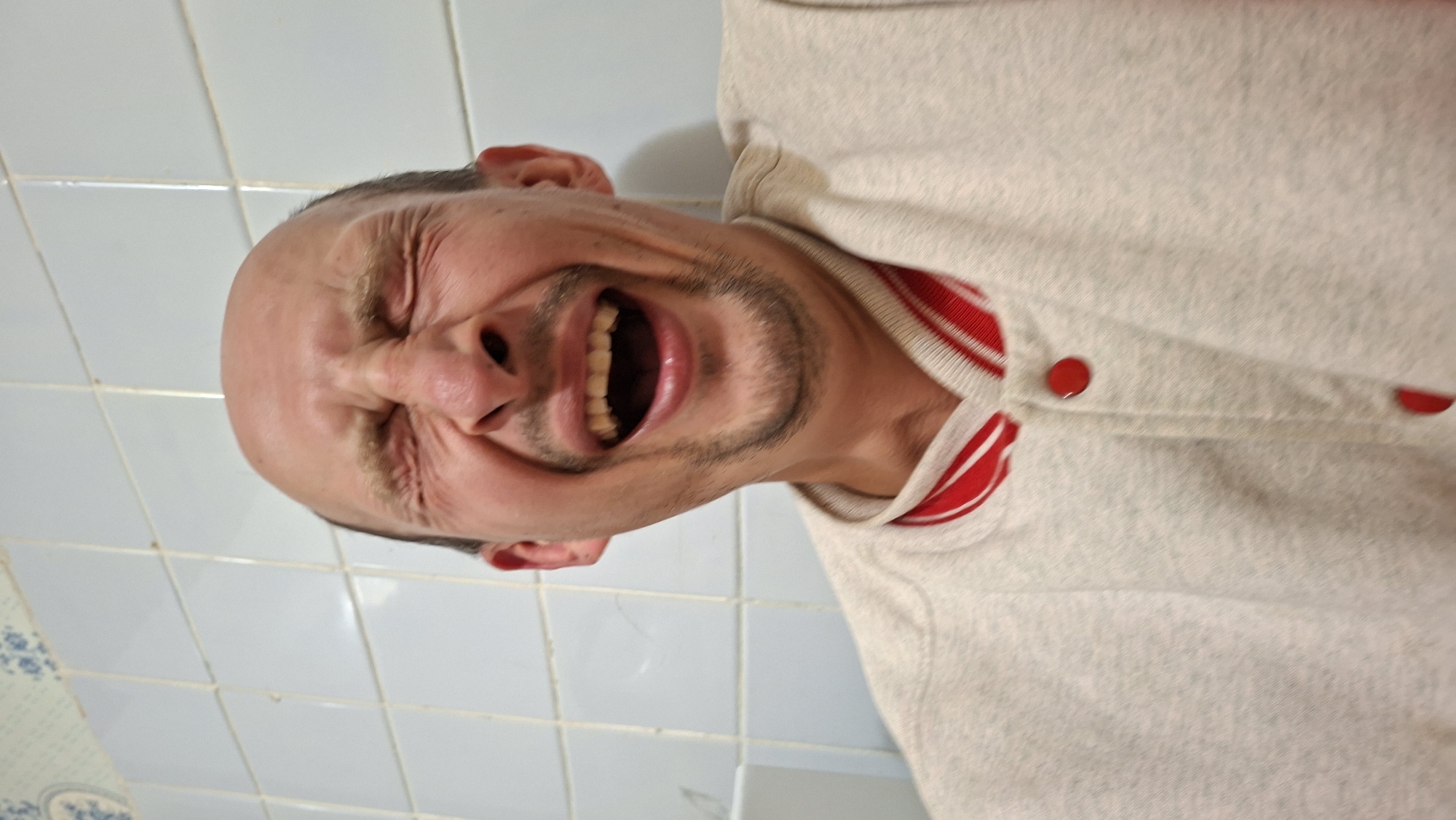 A man with a bald head is captured in a close-up, head-and-shoulders shot, laughing heartily. His eyes are squeezed shut, and his mouth is wide open, revealing his teeth. He has light brown eyebrows and a light beard stubble. He is wearing a light-colored jacket with red trim around the collar and red buttons down the front. The background consists of light blue tiled walls, typical of a bathroom or kitchen in a residential setting. The image is taken from a low angle, looking up at the man. There is no information available about the specific location in Забалоцкі сельскі Савет, Belarus.