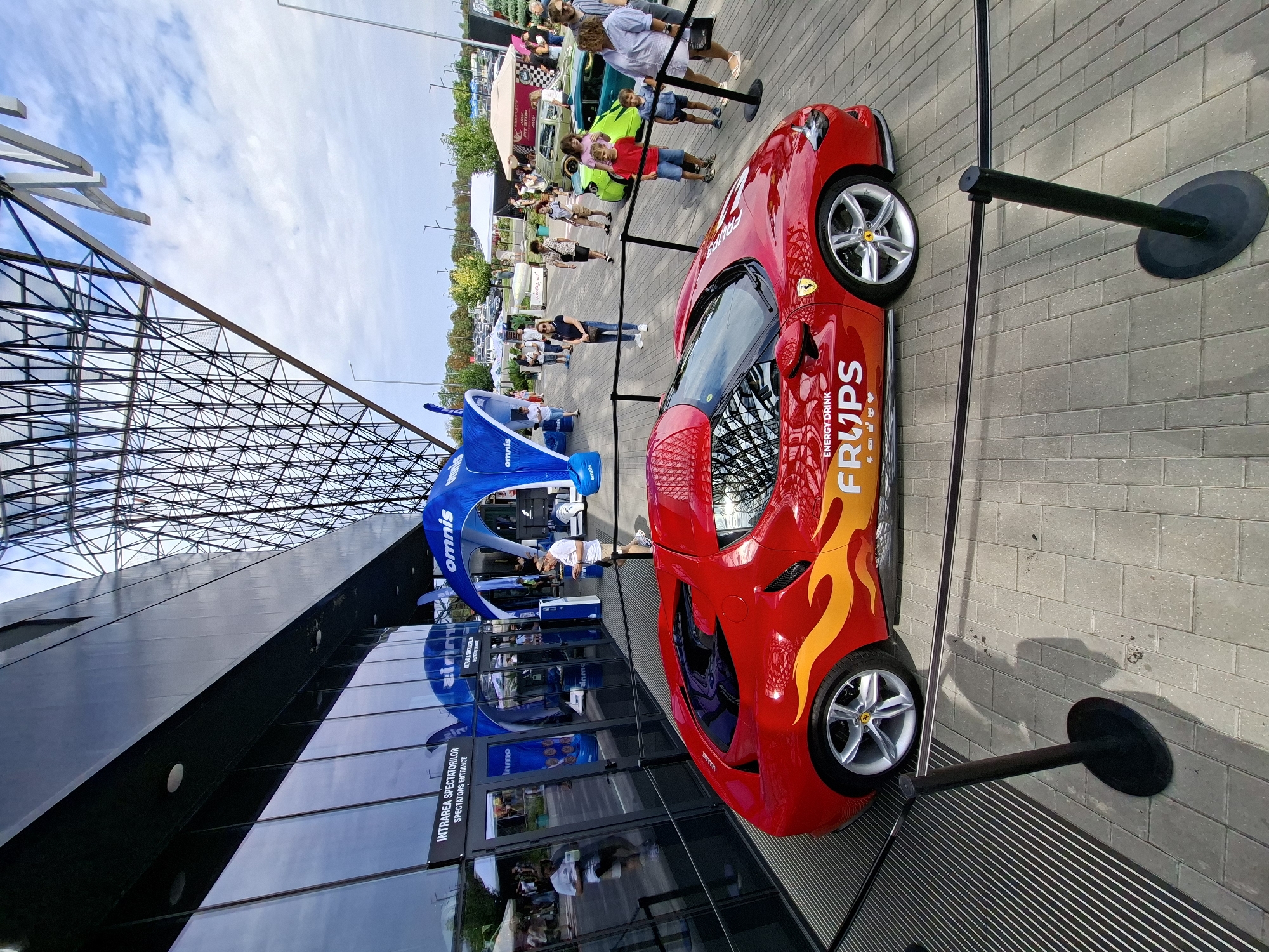 The image depicts an outdoor event on a paved walkway in front of a modern building with a glass facade. The building has a distinctive geometric metal structure extending upwards. Dominating the foreground is a vibrant red sports car, possibly a Ferrari, adorned with flame decals and prominently featuring the "FRUPS ENERGY DRINK" logo. Black velvet ropes and stanchions cordon off the vehicle, suggesting it is on display.
Behind the car, people are gathered, some looking at the car and others milling about. Several inflatable blue arches with the "OMNIS" logo are set up near the entrance of the building, which is labeled "INTRAREA SPECTATORILOR" (SPECTATORS ENTRANCE). Tents and other vehicles, including a light green sports car, are visible further in the background. The sky is partly cloudy, indicating it is daytime. This event appears to be a promotional activity or a car show taking place in the city of Stăuceni, Moldova, likely at or near a venue like the Stăuceni Arena or a similar event space.