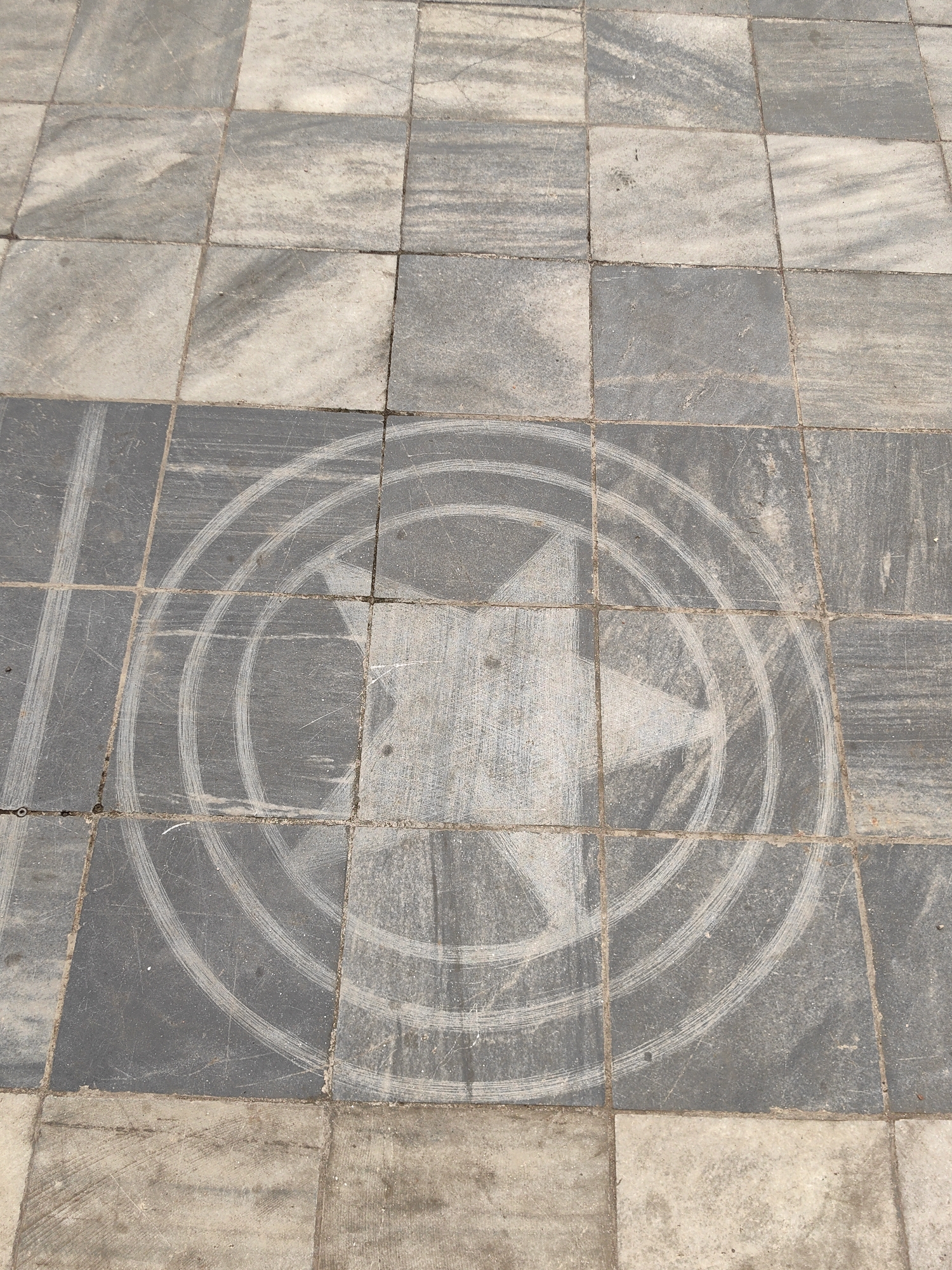 The image shows a tiled stone surface with a faint circular design containing a five-pointed star at the center. The tiles are gray and off-white, arranged in a grid pattern. The circular design, consisting of three concentric rings around the star, is lighter in color, as if it has been partially worn away or cleaned. The location is unknown.