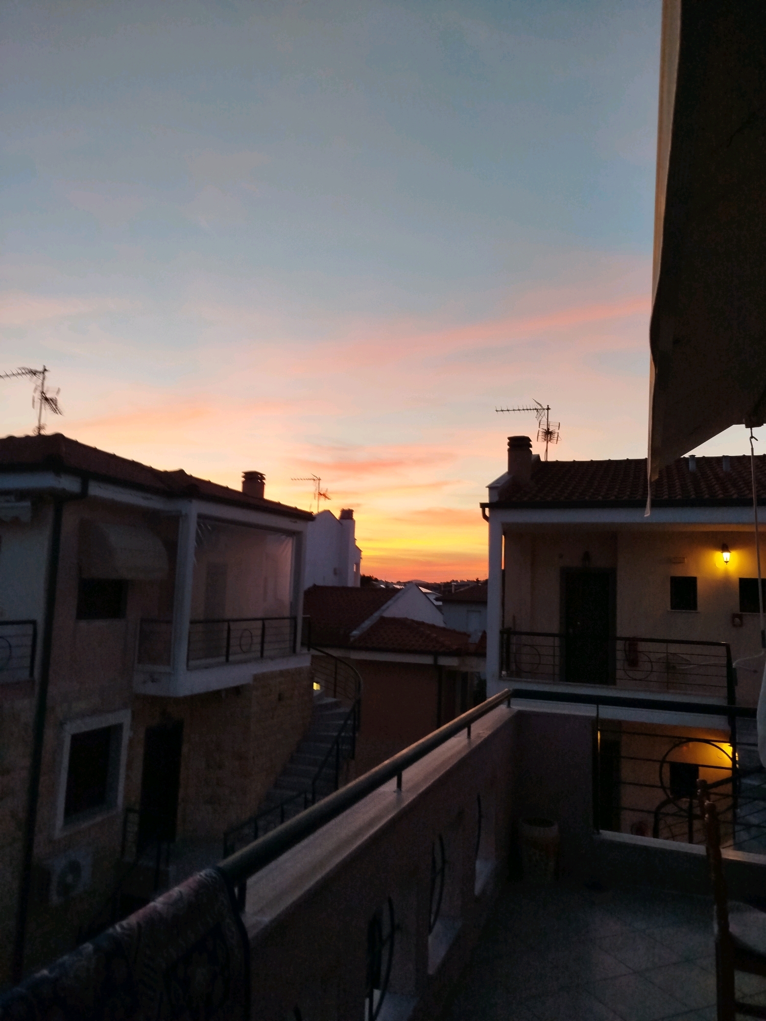 The image depicts a twilight scene from a balcony overlooking rooftops in the Sithonia Municipal Unit, Greece. The sky is a gradient of soft blues, pinks, and oranges, transitioning to yellow near the horizon. Several buildings with terracotta roofs are visible, some with balconies and windows. One building on the right has a lit lantern above its entrance. Antennas are mounted on some of the rooftops. The foreground shows a balcony railing with a patterned cloth draped over it. The overall atmosphere is calm and serene, characteristic of a Mediterranean evening.