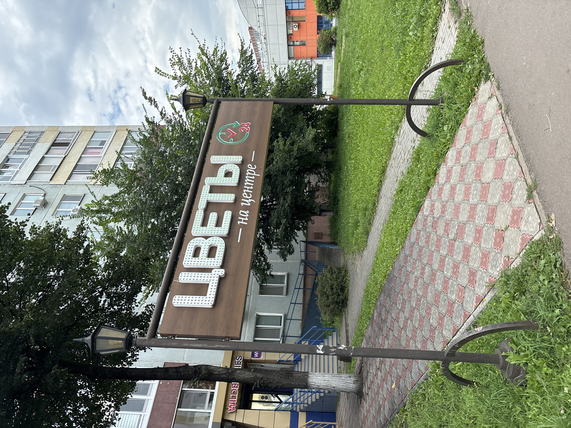 The image shows a street view in Naberezhnye Chelny, Russia, featuring a prominent wooden sign that reads "ЦВЕТЫ" (Flowers) in large, stylized white letters, followed by "на центре" (in the center) in smaller script. To the right of the text is a circular green logo with a checkmark and the number "24," indicating 24-hour operation. The sign is supported by two dark metal posts, each topped with an old-fashioned black lantern. The sign is positioned in front of a multi-story residential building with a light-colored facade and numerous windows. Adjacent to the building, a paved walkway with a red and gray checkerboard pattern leads towards the entrance. Lush green grass and trees surround the walkway and the base of the signposts. In the background, a modern orange and white building with blue windows is visible. The sky is overcast with clouds. No people are present in the frame.