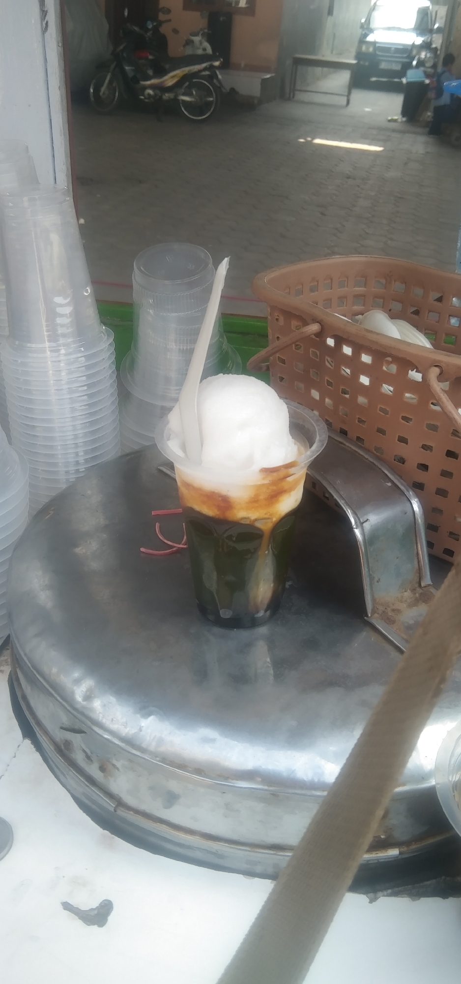 In Rahayu, Indonesia, a clear plastic cup filled with a layered beverage sits on a large, circular, metallic surface. The drink is topped with a white, scoop-like substance, possibly ice cream or shaved ice, and a white plastic spoon is inserted into it. Below the white topping, a dark brown liquid is visible, and beneath that, a dark green substance occupies the lower portion of the cup, with some small dark spheres at the very bottom. Red string or elastic bands are present near the base of the cup. To the left of the cup, stacks of clear plastic cups are arranged. To the right, a brown woven basket is visible, containing more white items, possibly more cups or food. In the background, a motorcycle is parked, and further back, a black vehicle is parked in an open area with a paved ground. The overall scene appears to be an outdoor vending stall or a street-side food service area.