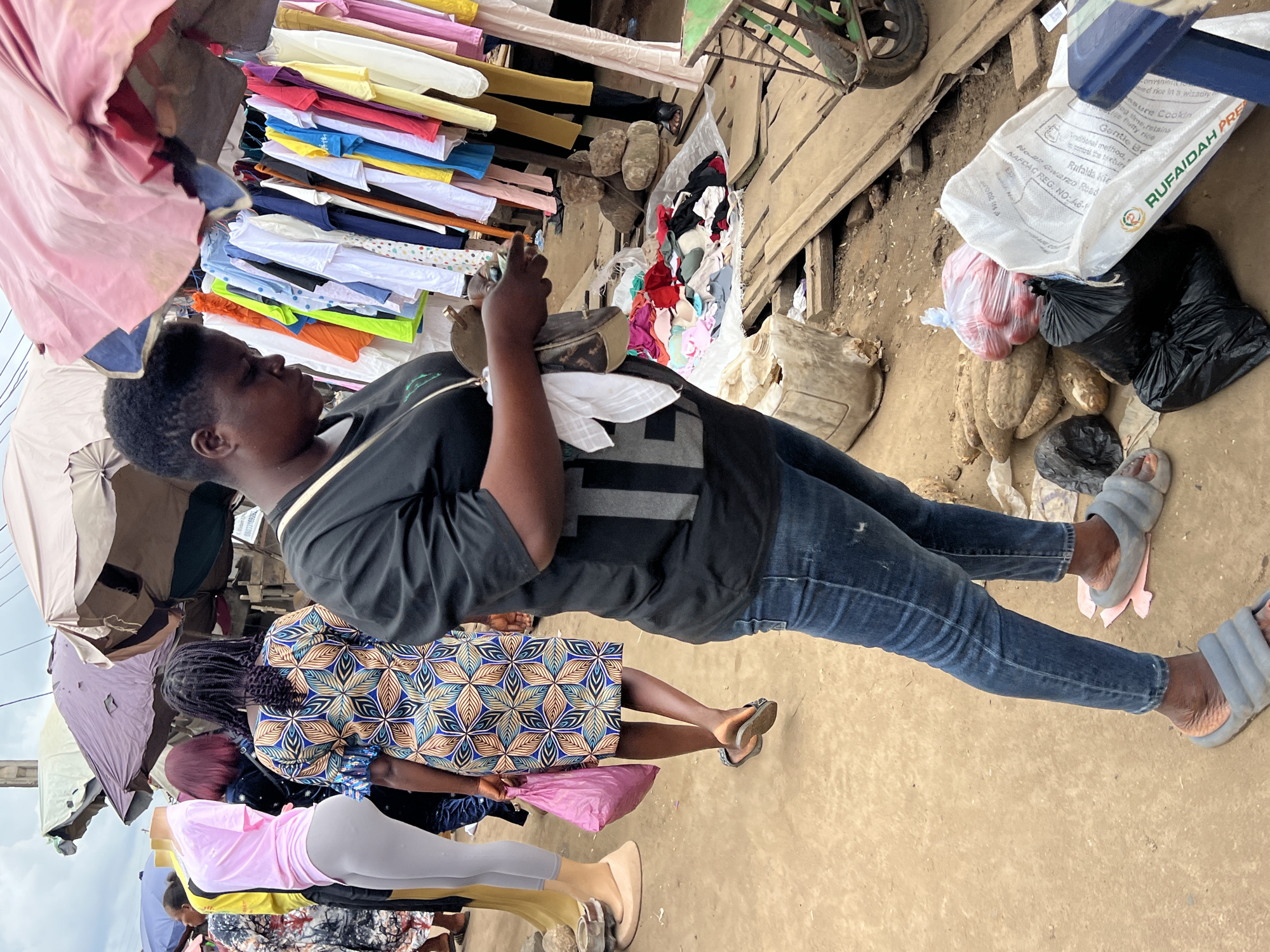This is a vertical, full-shot image taken outdoors, likely in a market in Asaba, Nigeria. The scene depicts several people and a variety of objects, including clothing and produce, suggesting a bustling marketplace environment.
In the foreground, a person is shown from the waist down, wearing blue jeans and grey sliders. They are holding a pink plastic bag in their right hand. To the left of this person, a mannequin dressed in a pink top and grey leggings stands upright, partially obscured by fabric. Behind the mannequin, another person is visible, also appearing to be a shopper or vendor.
Further into the image, a woman with dark braided hair is captured from the waist up, wearing a black t-shirt and blue jeans. She is looking down and appears to be interacting with or examining something in her hands. Next to her, a stall is laden with neatly folded clothes in various colors, including white, yellow, pink, red, and blue. Some of these are t-shirts, and others are patterned fabrics.
The background reveals more market elements. A large white sack with text, including "RUFAIDAH PRE" and other illegible markings, is prominent, lying on the ground next to a black plastic bag and a pile of root vegetables. Other assorted items, including more bags and what appear to be umbrellas, are scattered around. The ground is a mix of dirt and sand. The overall impression is of a typical vibrant and busy open-air market, possibly in the mid-morning or afternoon given the lighting.