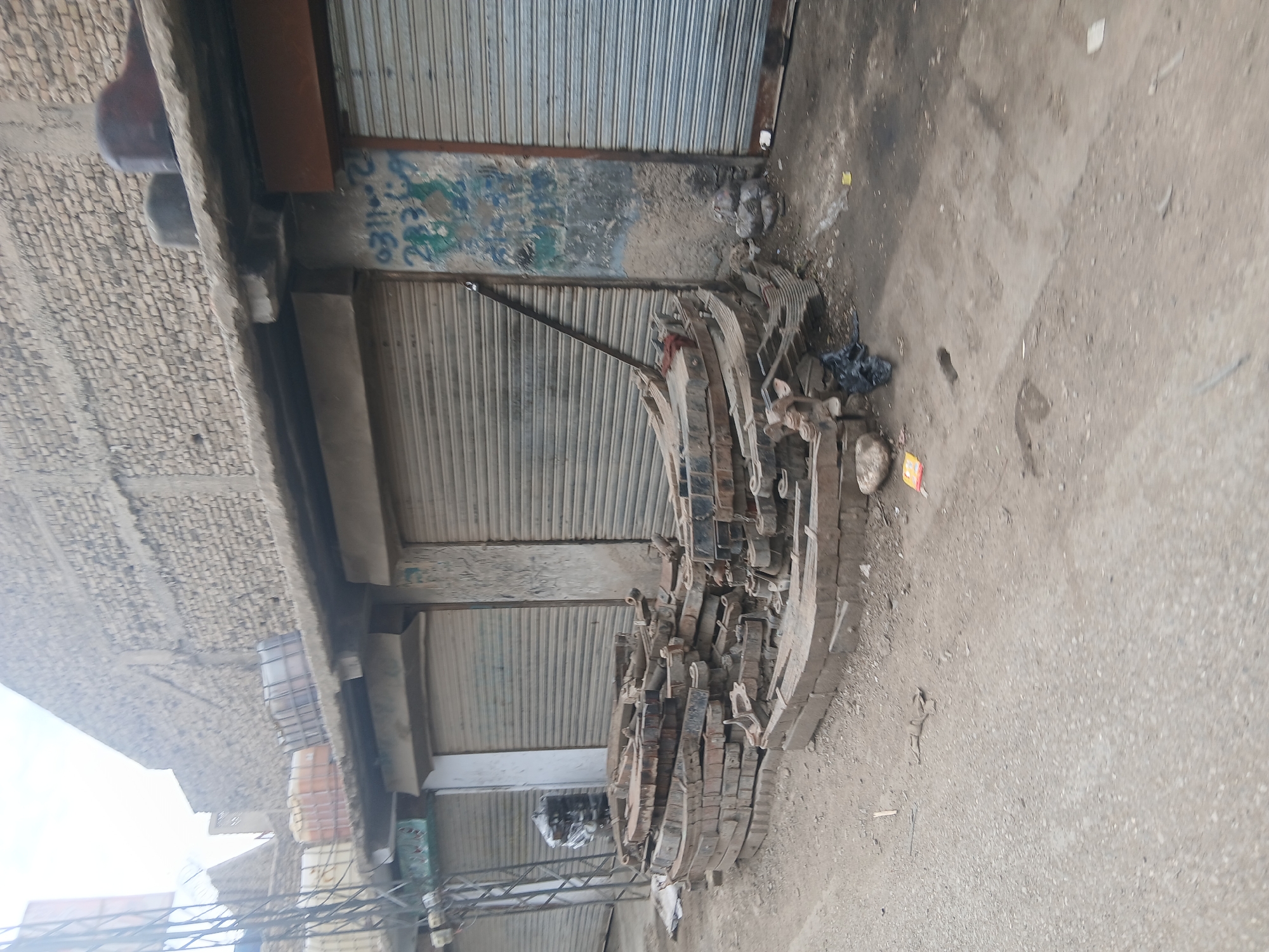The scene depicts a street in Quetta City Tehsil, Pakistan, lined with buildings featuring closed, corrugated metal shutters. A large pile of used vehicle leaf springs lies on the ground near the buildings. The street surface is concrete or asphalt and appears worn. No people are visible. The environment appears urban and somewhat dilapidated.
