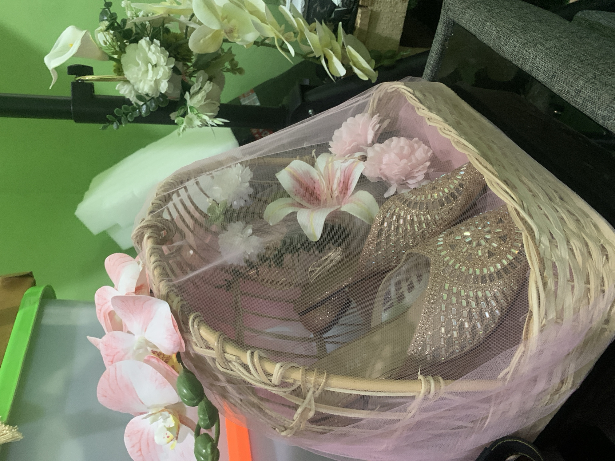 The image shows a wicker basket filled with decorative items, including a pair of gold, embellished flat shoes, several artificial flowers, and a pink tulle fabric draped over the contents. The basket is placed on a dark surface, possibly a table, in front of a grey upholstered chair. In the background, there are glimpses of green walls, artificial floral arrangements, and a black stand. The overall presentation suggests a gift or display for a special occasion. The location is not specified in the image, and therefore cannot be identified as Kuala Pembuang, Indonesia.