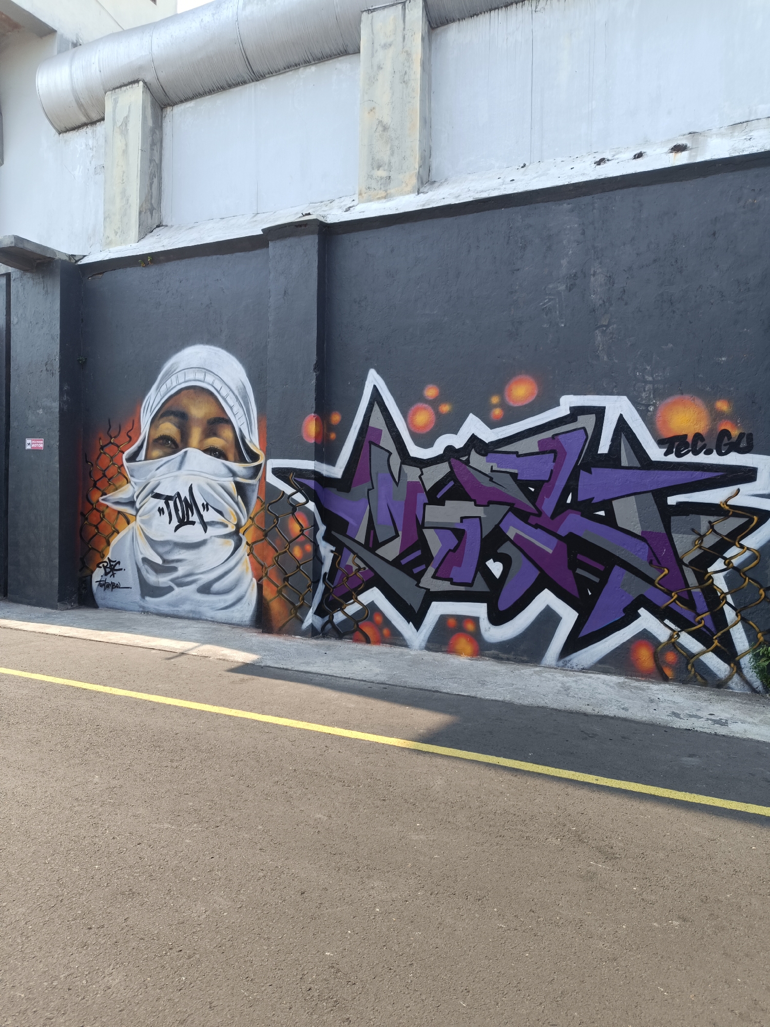 The image captures a section of an outdoor wall adorned with graffiti, located in Yogyakarta, Indonesia. On the left, a realistic mural depicts a person's face, with their eyes visible above a scarf covering their nose and mouth. The word "TOM" is prominently written in black, stylized lettering on the scarf. The mural is set against a dark grey wall with orange circular accents and a chain-link fence in front of it. To the right of the mural, a vibrant graffiti tag in purple, grey, and white fills a large portion of the wall, with "TEC.GU" written in black above it. The wall is part of a building's exterior, with a light-colored upper structure and pipes visible. In the foreground, a paved surface, likely a road or sidewalk, features a single yellow line running parallel to the wall. The scene appears to be illuminated by natural sunlight, casting shadows on the ground.