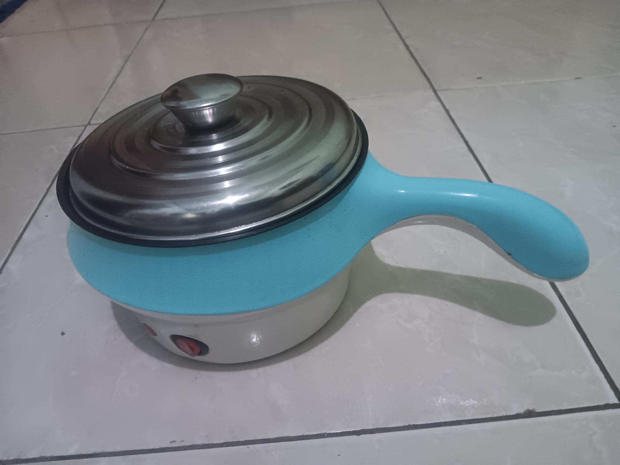 A light blue and white electric cooking pot with a silver lid is placed on a tiled floor. The pot has a light blue handle and two red buttons on its base. The floor tiles are a light gray color with darker grout lines. The scene appears to be indoors.