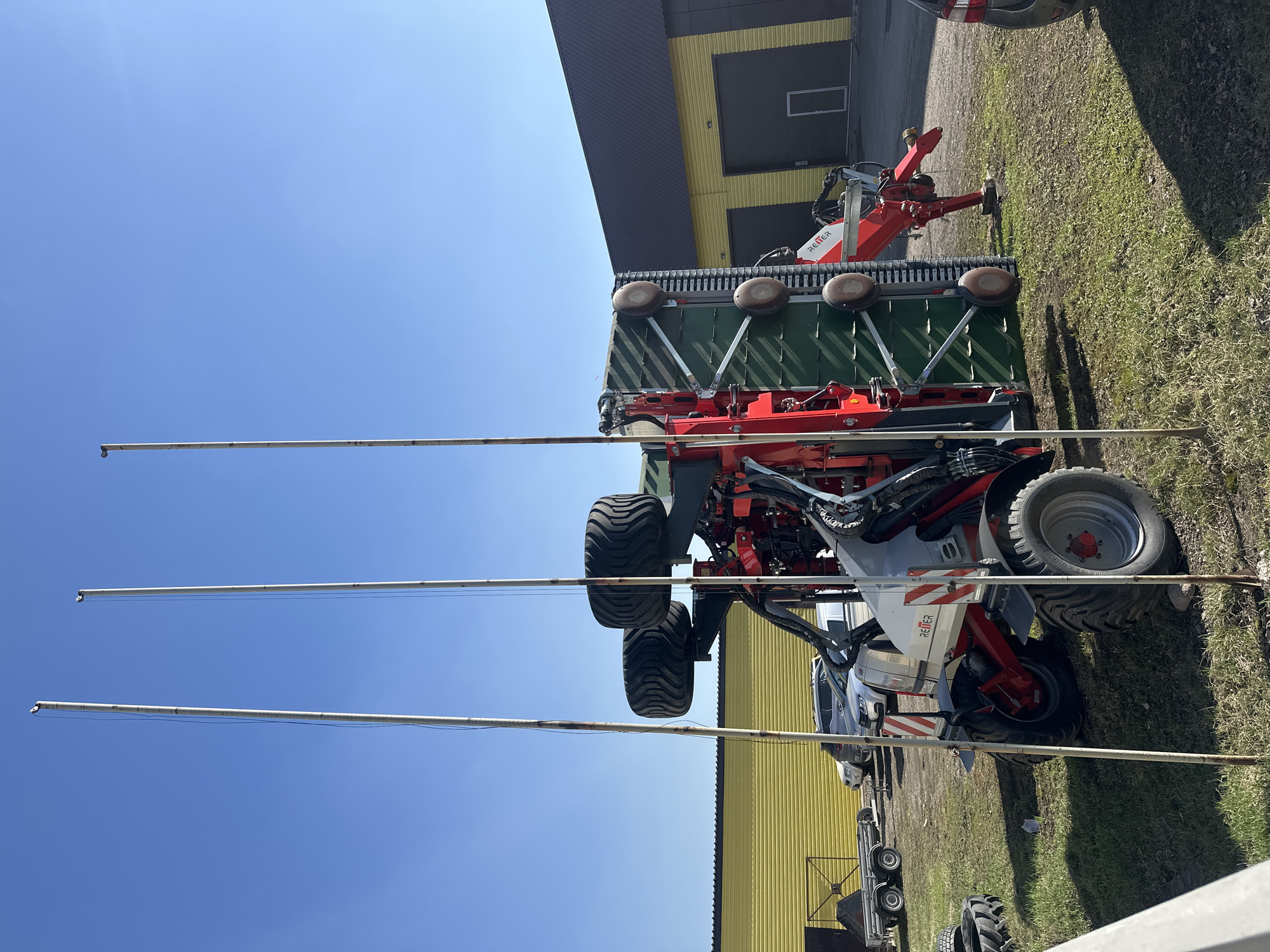 The image, correctly oriented vertically, presents a scene dominated by large agricultural machinery parked outdoors. In the foreground, a substantial self-propelled machine, likely a forage harvester or sprayer, with large, robust tires, displays a white, grey, and red color scheme. Attached to its front is a wide, green and red agricultural implement, characterized by a series of large, brown, disc-shaped rollers or wheels mounted along its upper edge, and a slatted or ridged green body.
Three thin, metallic poles extend vertically across the left side of the frame, partially obscuring the view of the machinery and the sky.
In the mid-ground, to the right of the main machinery, another red agricultural machine is visible, with the brand name "REITER" clearly printed in white on its side. Further in the background, a large, modern industrial building with a dark grey roof and prominent yellow wall paneling stretches across the left half of the image. The building features several dark, rectangular openings, possibly garage doors. The ground is covered in grass, which appears somewhat dry in places.
The scene unfolds under a clear, bright blue sky, indicating sunny weather. No human activity is depicted; the machines appear to be stationary. The setting suggests an agricultural facility, farmyard, or a machinery dealership in Biysk, Russia.