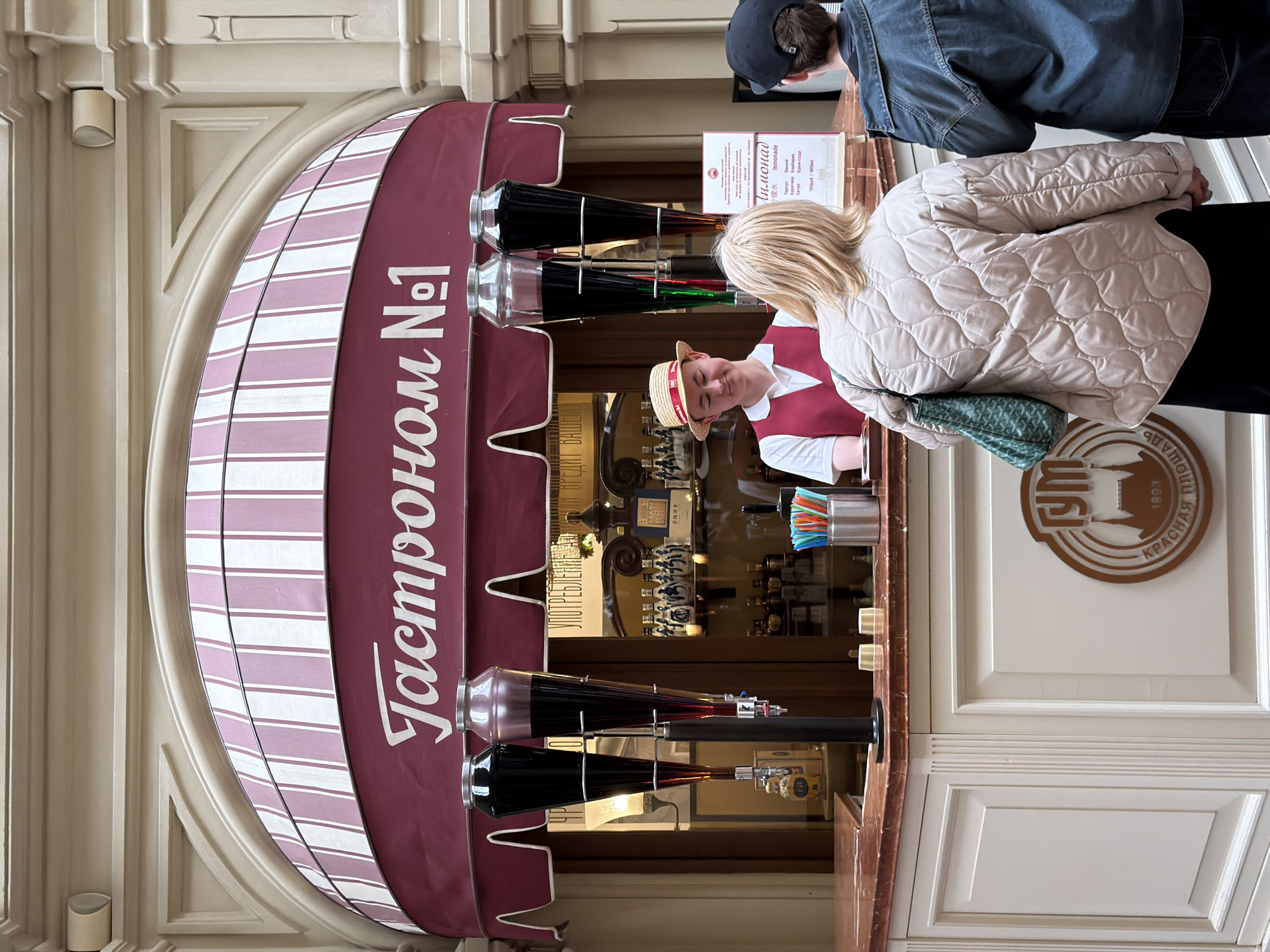 The image shows the "Gastronom No. 1" lemonade stand, which sells lemonade. The stall has a maroon and white striped awning, with the words "Gastronom No. 1" written in white Cyrillic letters. There are glass containers of beverages being dispensed. The signage is in Russian and Chinese. A blonde woman is standing at the counter and another person is visible in the upper right. A sign in the background indicates that this stall is located in the GUM department store on Red Square in Moscow.