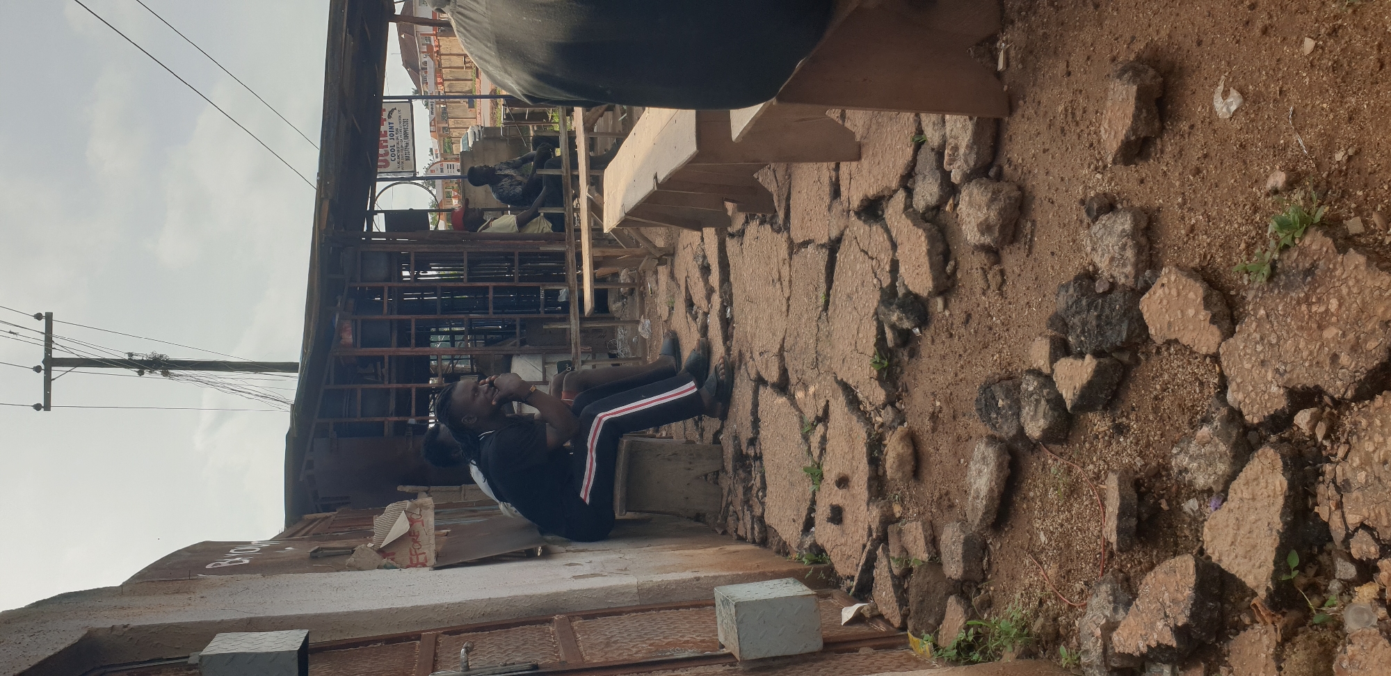 The image depicts an outdoor scene in Yelwa, Nigeria, under an overcast sky. The foreground features a severely damaged and uneven concrete pavement, fragmented into numerous pieces with reddish-brown soil and small stones visible between cracks. Patches of small green vegetation grow through the ground.
On the left, a weathered building with a corrugated metal door is visible. A white concrete block rests at its base. Faint, white lettering "BAYO" is visible on the wall. A small, torn cardboard sign is affixed to the wall, bearing the partially legible text "BEFORE 12". A dark, small object rests on the broken pavement near the wall.
In the mid-ground, a man with dreadlocks, wearing a black t-shirt with red and white striped sleeves, black trousers with vertical red and white stripes, and black sandals, sits on a low, rustic wooden bench. He is looking towards the right, with his right hand near his chin. Partially obscured behind him is another man also sitting on the same bench.
Further to the right, a rudimentary open-air structure with a metal frame and corrugated roof stands. Inside this structure, a white rectangular sign for "UCHE COOL JOINT" is prominently displayed, listing an address "1. Umu Junction Off Taiwo Road, FCE, Okene, Kogi State" and two phone numbers "08033784661, 08099157203". Several men are seated on simple wooden benches within and around this structure; one wears a red cap, another a patterned shirt. A long, unpainted wooden bench extends from this area towards the foreground right. The back of a person in a dark shirt is partially visible in the extreme foreground right.
In the upper central portion of the image, a wooden utility pole with multiple entangled wires extends vertically against the cloudy sky. In the far background, beyond the informal structures, residential buildings with red roofs are visible. A thin red wire and scattered small white debris are present on the ground.