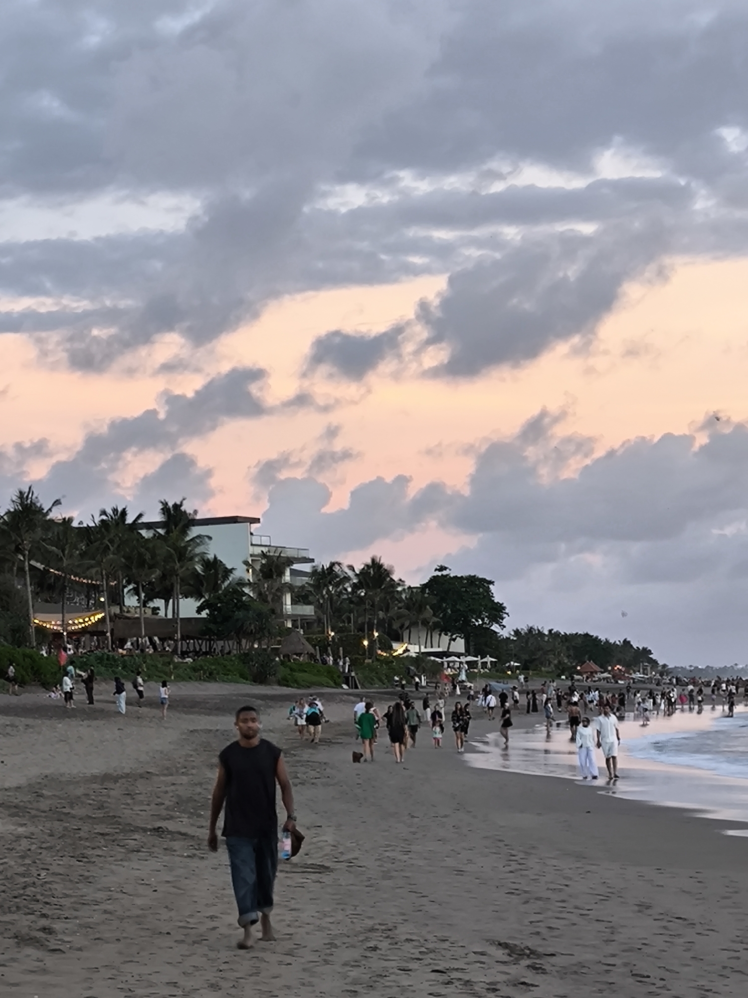 The image captures a beach scene in North Kuta, Indonesia, at dusk. The sky is filled with clouds tinged with pink from the setting sun. Numerous people are walking along the shoreline, some closer to the water's edge where waves are visible, and others further back on the sand. In the foreground, a man is walking towards the camera, carrying a water bottle and a pair of shoes. Palm trees and buildings can be seen in the background, along with string lights, suggesting a restaurant or entertainment venue is nearby. The beach has dark sand.