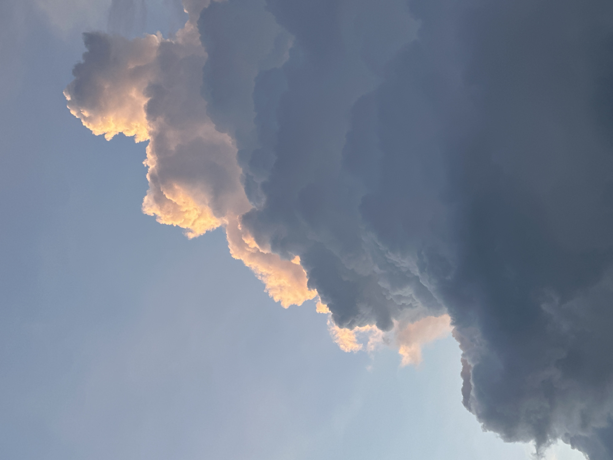 The image shows a low-angle view of clouds in Datça, Turkey. The sky is predominantly blue, transitioning into shades of light gray and white near the horizon. A large, layered cloud formation occupies a significant portion of the image. One section of the cloud is illuminated by the sun, giving it a bright yellow-orange glow along its edges and contours, while the other section appears darker, varying in shades of gray and blue, suggesting a storm cloud. There are no people or objects visible other than the sky and clouds.
