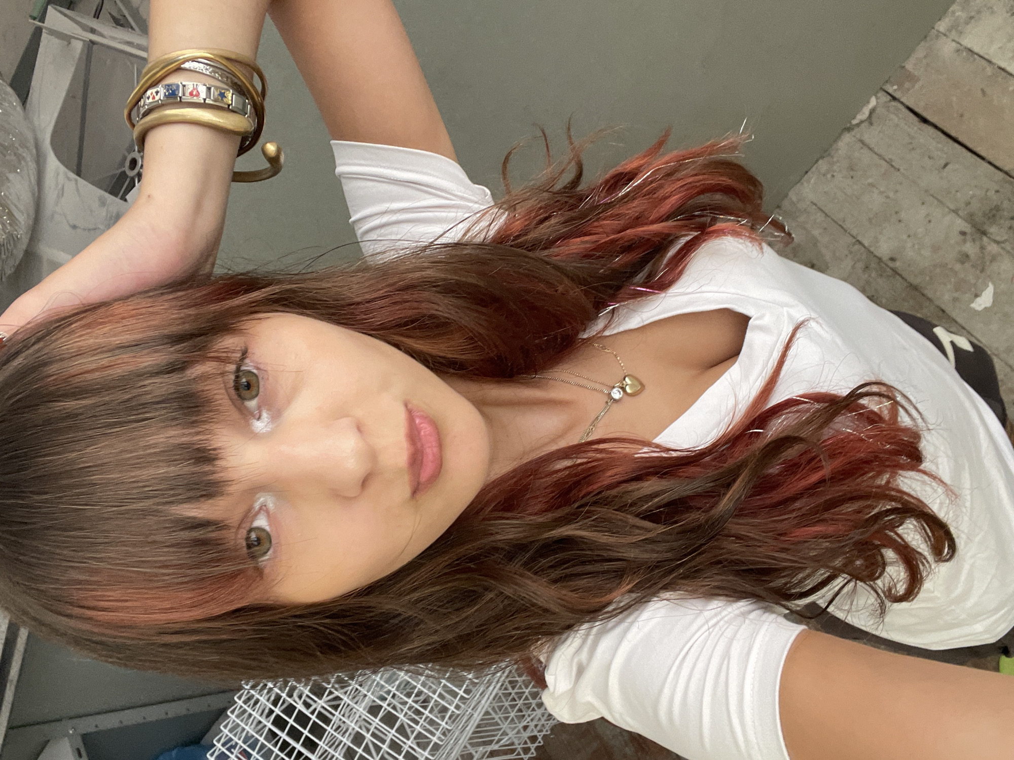 A young woman with long, dark, wavy hair with streaks of red and silver tinsel is posing for a selfie. She is wearing a white V-neck t-shirt and several bracelets on her left arm. Her right hand is raised to the side of her head. She is wearing a necklace with a heart-shaped pendant. The background consists of a white wire rack and a gray wall, with wooden floorboards in the foreground. The image appears to be taken indoors, possibly in an unfinished or construction area. The presence of a wire rack and the unfinished flooring suggest a storage area or a space undergoing renovation. The overall lighting is natural, indicating an indoor setting with natural light sources. There is no discernible indication of the city of Raanana, Israel.