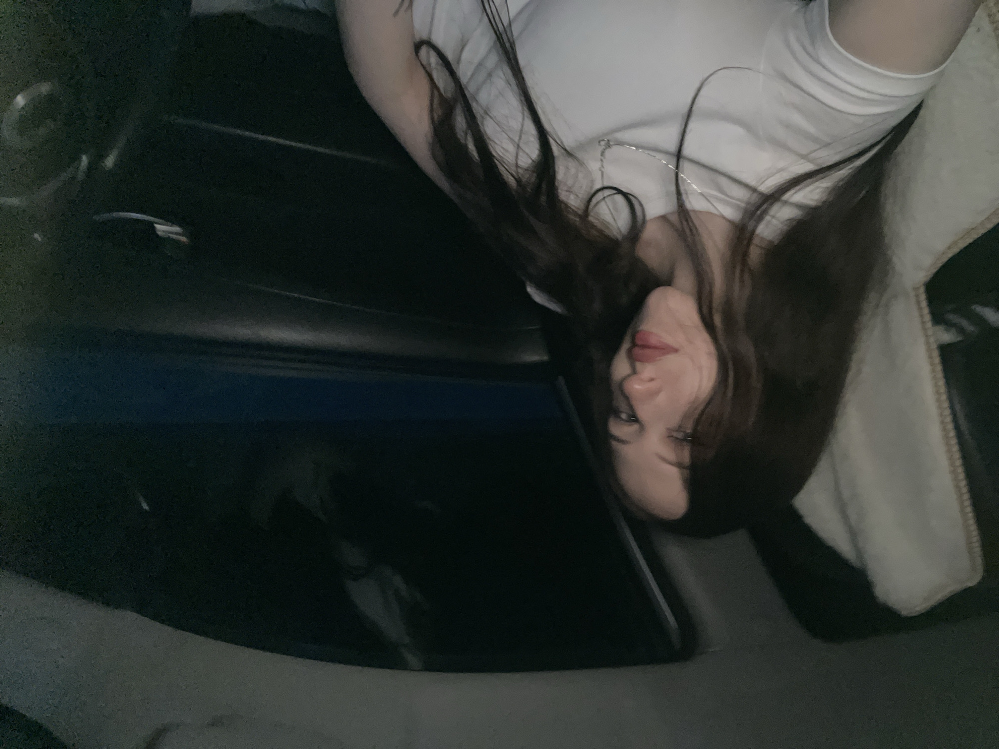 A young woman with long, dark hair and fair skin is shown from the chest up, sitting in the passenger seat of a car at night. She is wearing a white t-shirt and a silver necklace. Her head is tilted slightly, and she is looking towards the camera with a neutral expression. The interior of the car is dark, with the window behind her reflecting a dark, blurry image, possibly of buildings or lights. The seat next to her is covered with a cream-colored fluffy material. The image appears to have been taken with a low-resolution camera, resulting in a grainy texture. The lighting suggests an urban environment at night, with the car likely parked on a street in Gavrilovka, Russia.