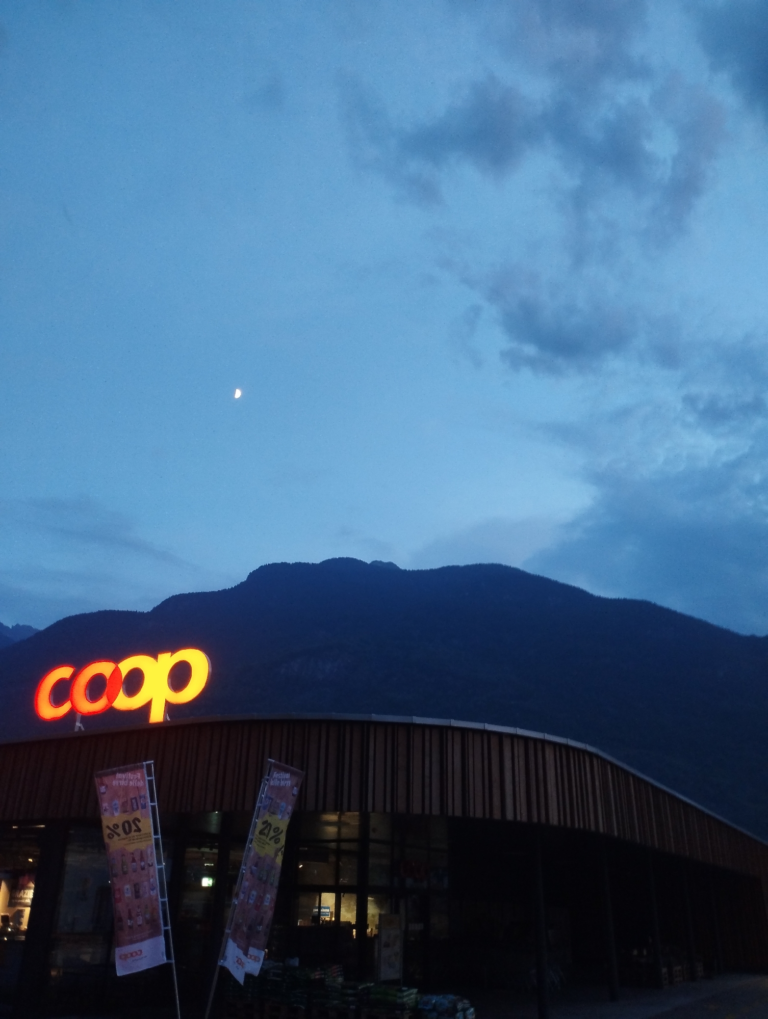 An exterior, low-angle, full shot shows a Coop grocery store in the foreground, with the store's illuminated "coop" logo prominently displayed above the entrance. The building has a modern design with vertical wooden slats. Two promotional banners are placed in front of the store, one displaying "20%" and the other "21%" along with product images. In the background, dark, silhouetted mountains rise against a twilight sky. A crescent moon is visible in the upper left portion of the sky. The lighting suggests it is dusk or early evening. There are no people visible in the scene.