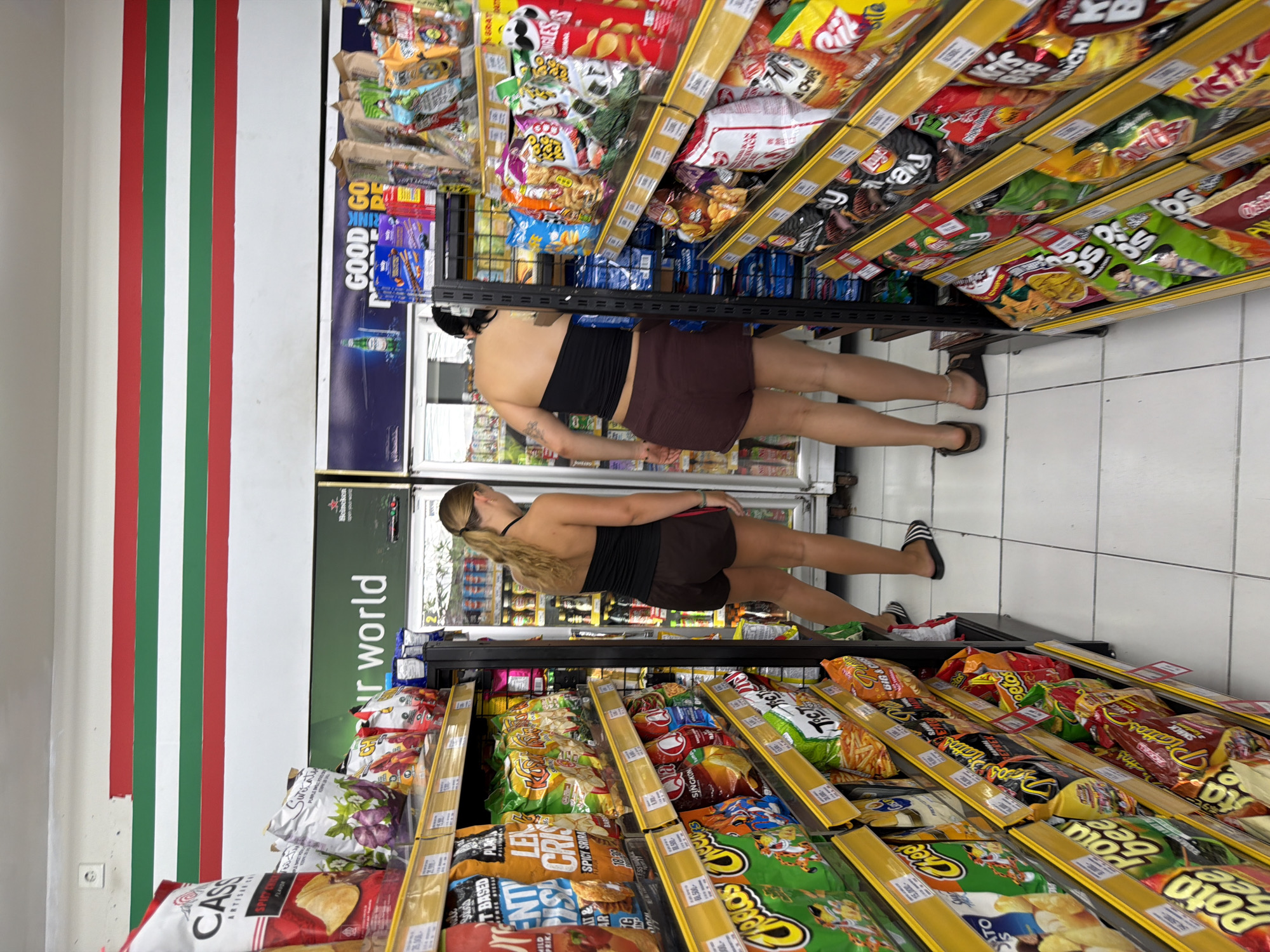 In a convenience store in North Kuta, Indonesia, two women are browsing the snack aisles. The store has brightly lit shelves stocked with various chips, cookies, and drinks. The walls are painted white with horizontal red, white, and green stripes near the ceiling. The floor is tiled in white.
The woman on the left, with blonde hair tied in a ponytail, wears a black halter top and brown shorts. She is looking at the products on a shelf.
The woman on the right, with dark hair, is wearing a black strapless top and dark brown shorts. She is bent over and appears to be reaching for something on a lower shelf. Her left arm is visible and has a tattoo on it.
In the background, refrigerators are filled with drinks. Various brands of snacks are visible on the shelves, including Lay's, Cheetos, and Pringles.