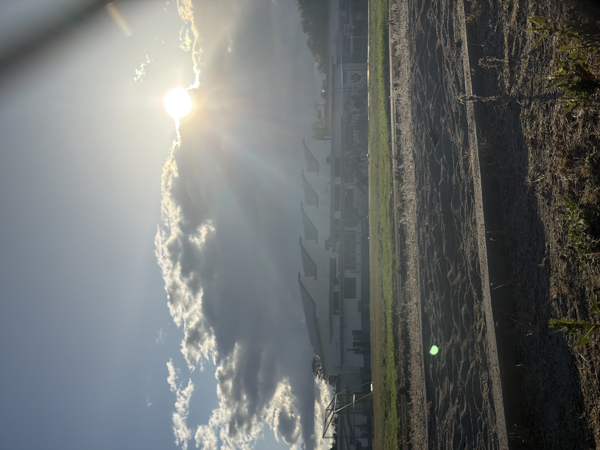 The image shows a landscape under bright sunlight, likely in Burbank, United States of America. A line of white buildings, probably apartments or offices, stretches horizontally across the midground of the image. Clouds obscure part of the buildings and the sun, which shines brightly in the upper left corner. The foreground consists of a dirt or sandy area with some vegetation along the right side. Tire tracks or grooves mark the dirt surface. A grassy strip separates the buildings from the dirt area. The scene is oriented in a way that makes the horizon appear vertical in the frame.
